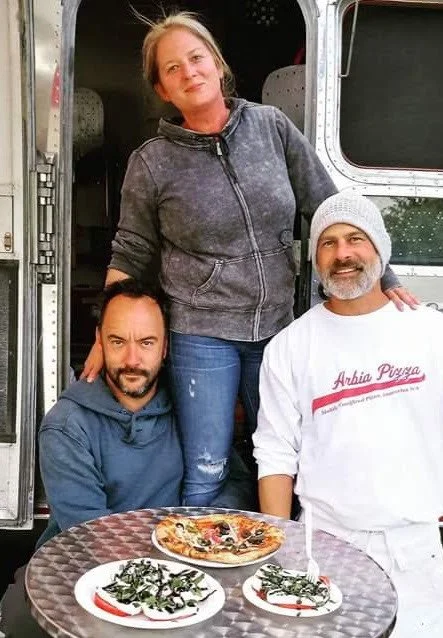 Three people, two men (one being Dave Matthew of the Dave Matthews Band) and one woman, posing outside a food truck with two pizzas and salad on a table in front of them. 
