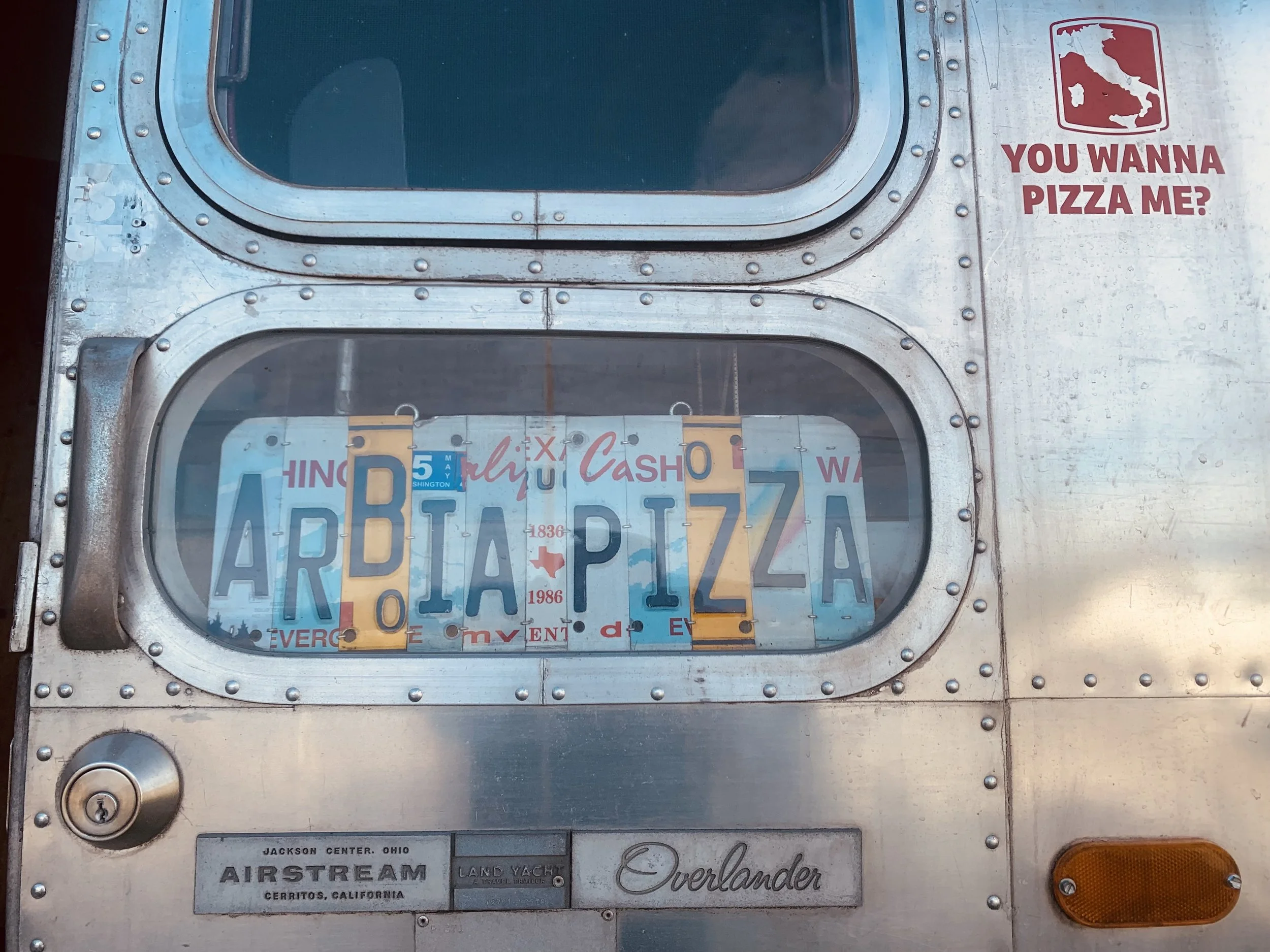 Close-up of a vintage metal trailer with a small window, stickers spelling Arbia Pizza, a mobile Pizza Trailer
