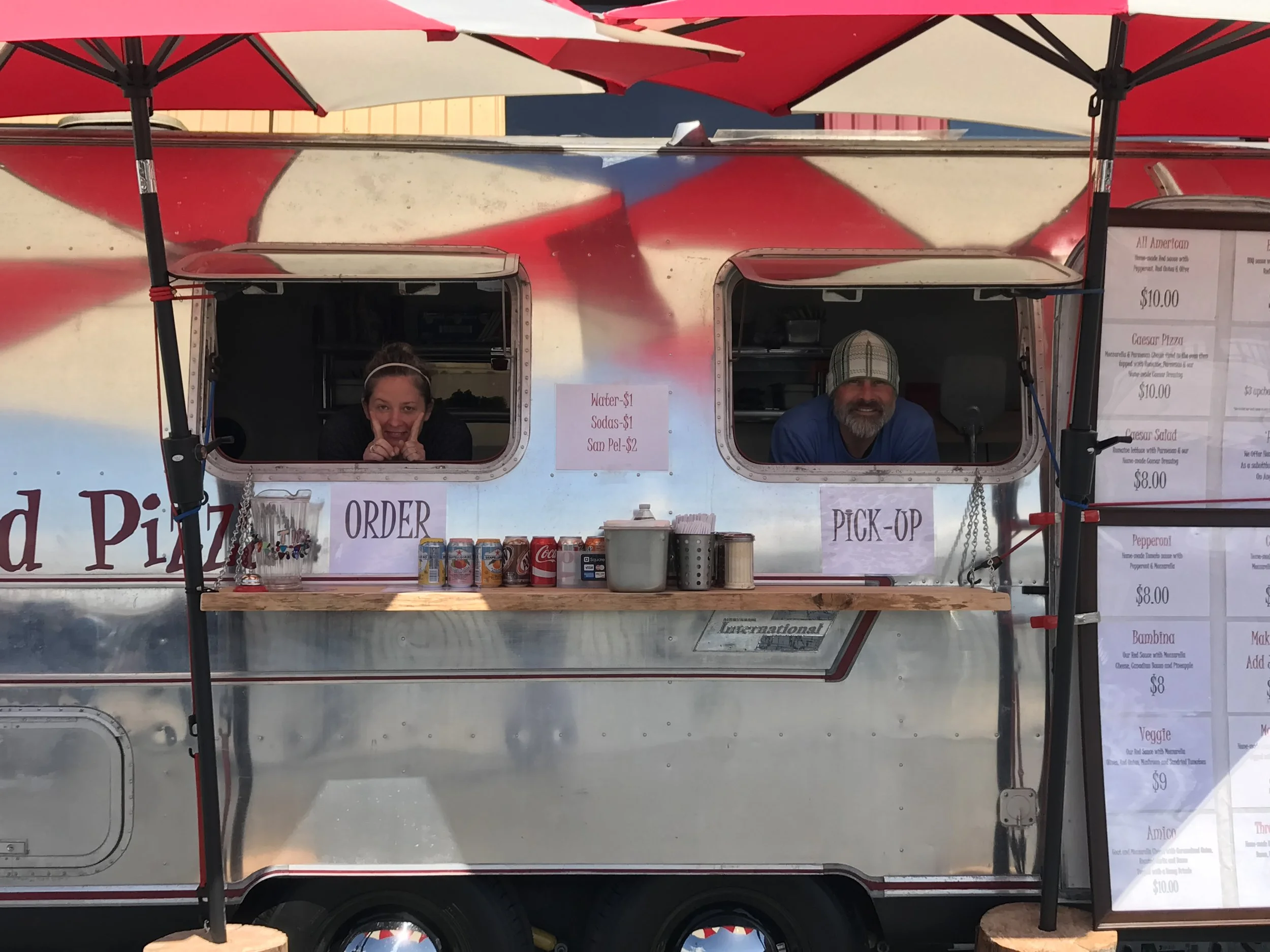 A food truck with two people inside, a woman on the left and a man on the right, smiling. The truck has a red and white striped umbrella on top. There is a menu at the side with various food options and prices, and cans of soda on the counter.