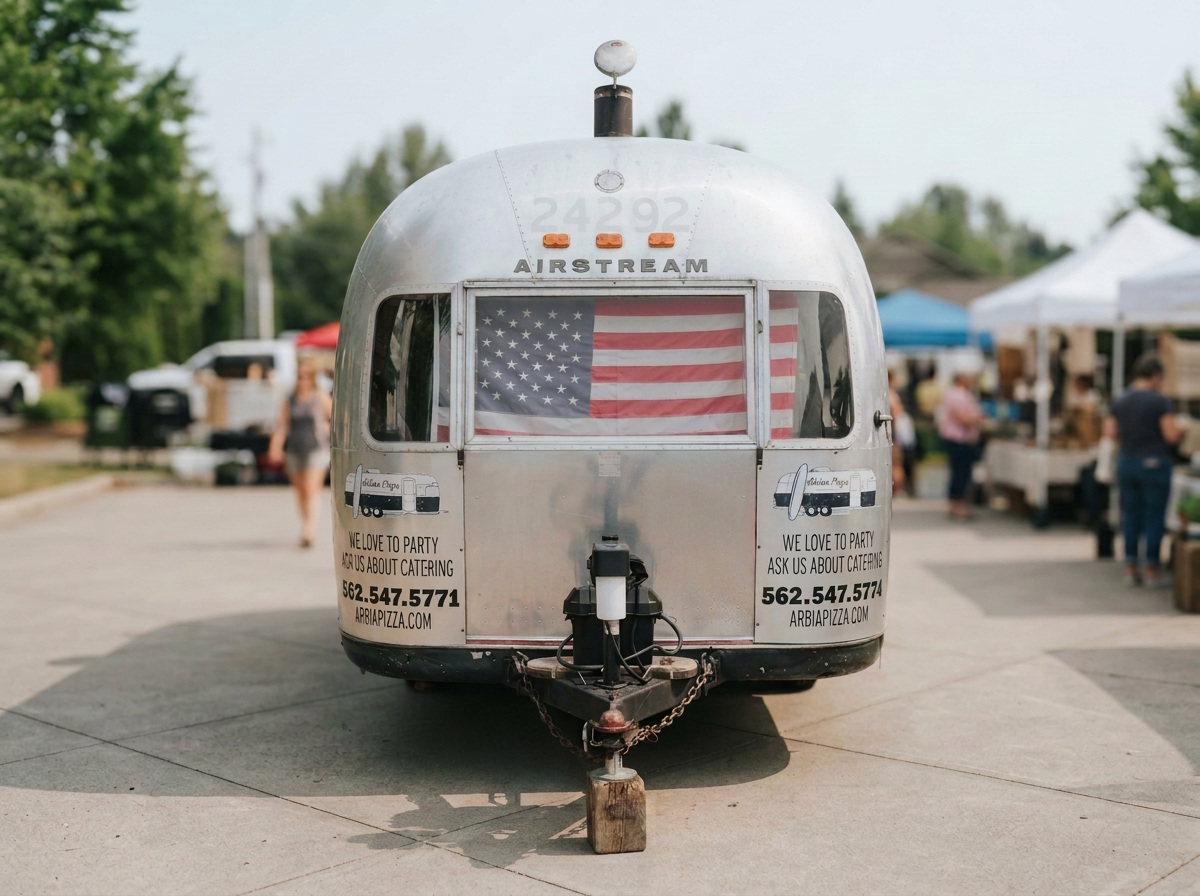 A silver vintage Airstream trailer with an American flag in the window, parked at an outdoor market with tents and people in the background.
