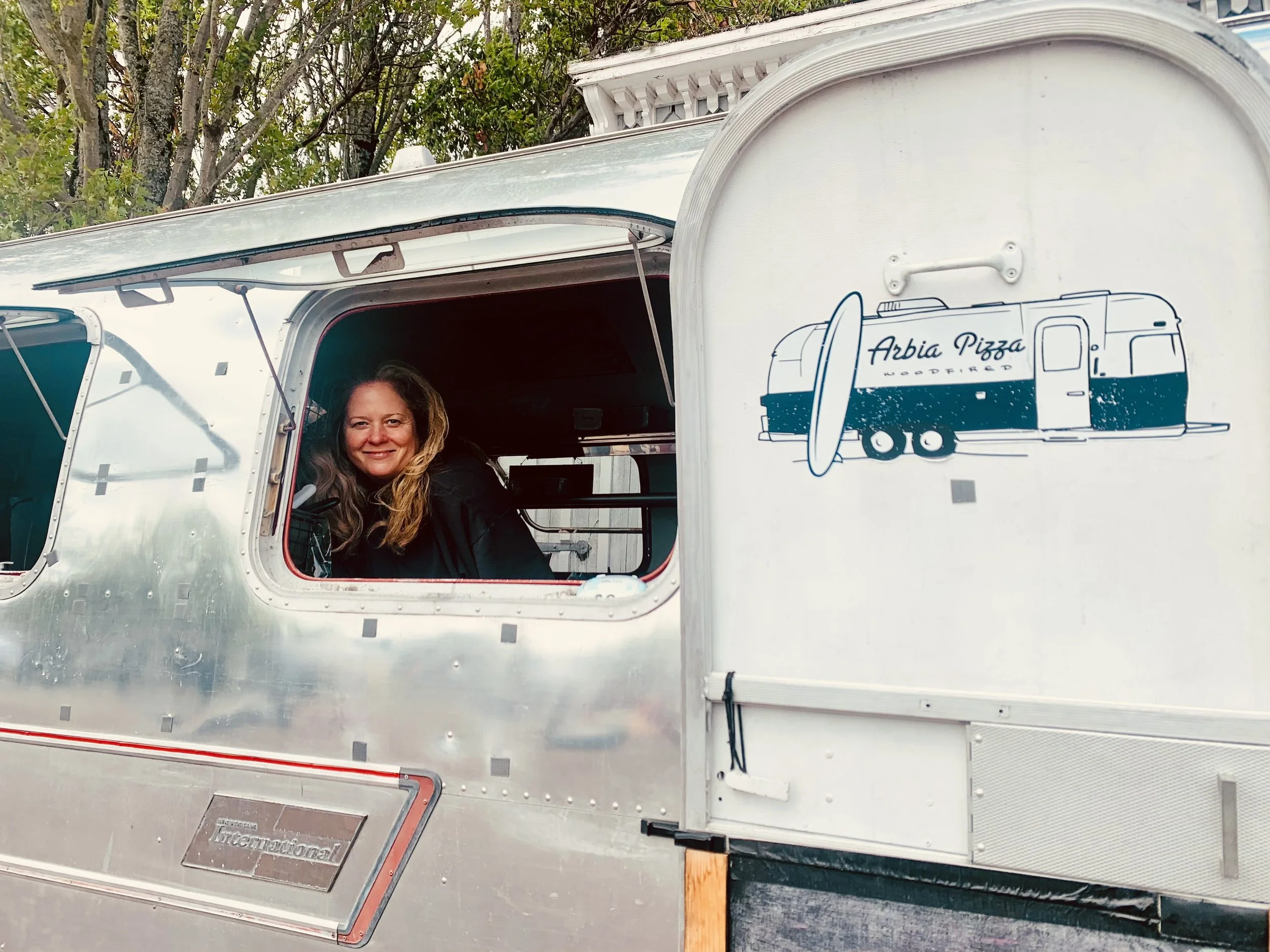 Person smiling and sitting inside a vintage Airstream trailer with a sign for Arbia Pizza on the exterior.