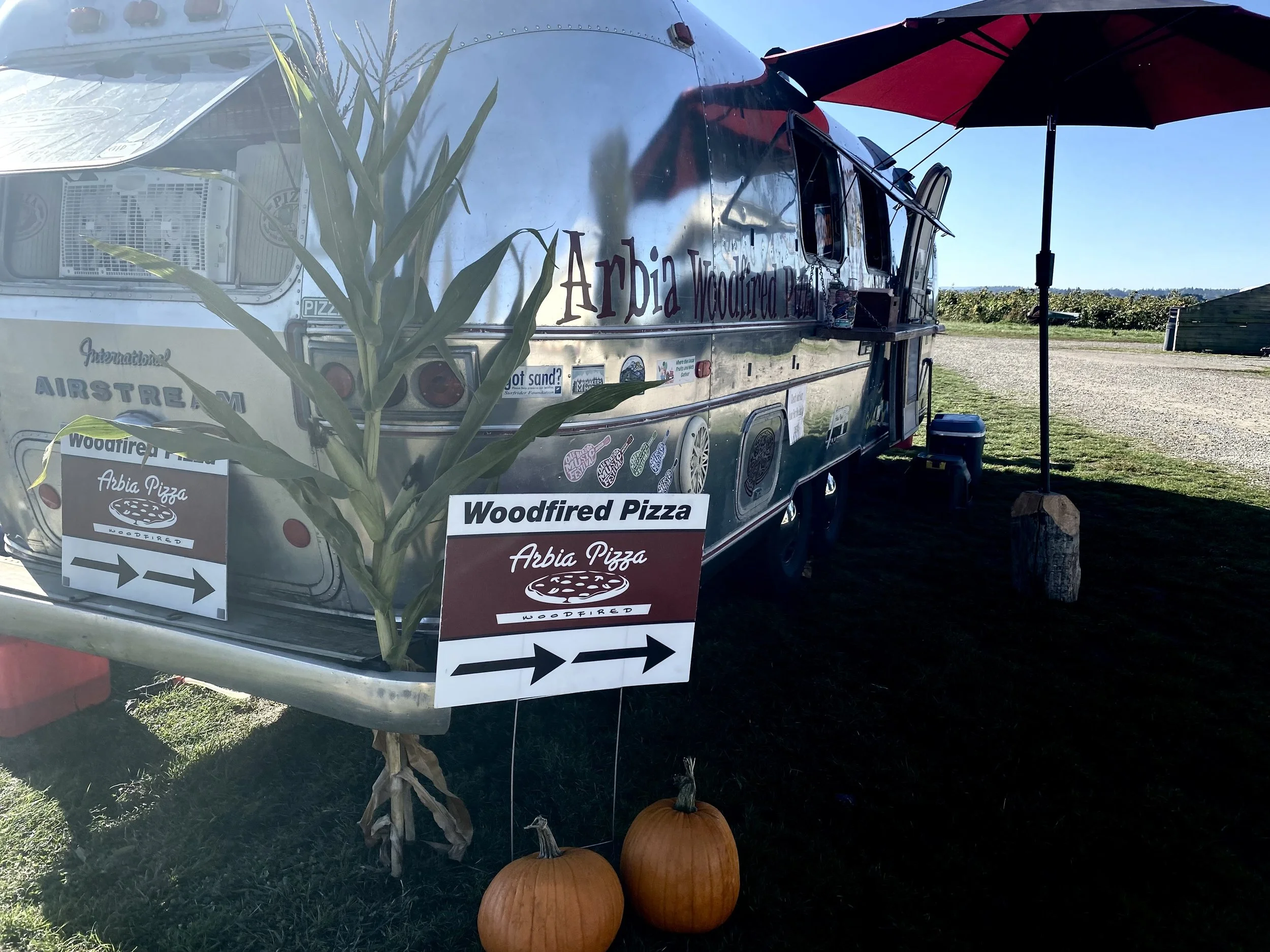 A food truck with a sign that reads 'Woodfired Pizza' parked outdoors with pumpkins in front and an umbrella attached. There is a plant in the foreground and the background shows a clear sky and distant fields.