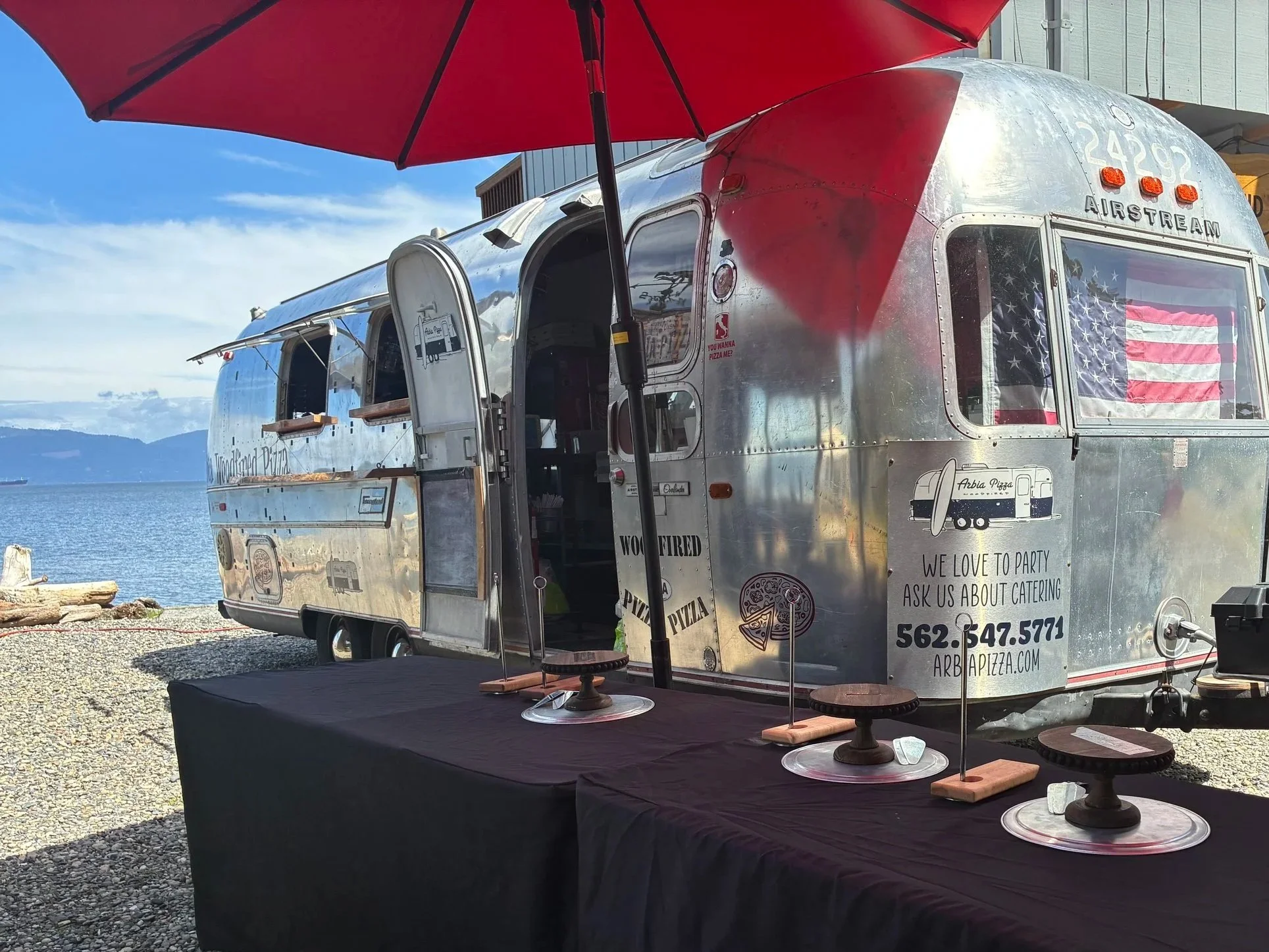 A shiny, metal Airstream food truck decorated with an American flag and patriotic star pattern, parked outdoors near a body of water with mountains in the background. There is a large red umbrella providing shade over a table in front of the truck.