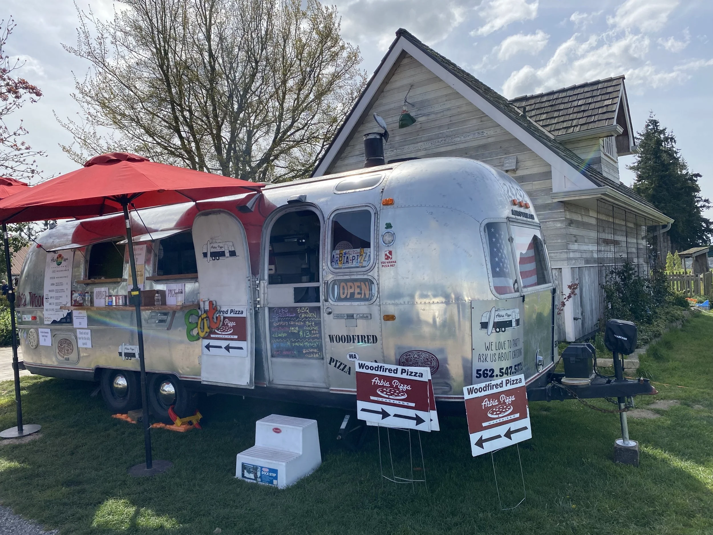 A silver vintage trailer converted into a pizza food stand with red umbrellas providing shade. Signage and menu boards are displayed outside, promoting Woodfired Pizza. The trailer has signs with contact information and promotional messages. The setting appears to be a backyard or small outdoor event space, with trees and a house in the background under a partly cloudy sky.