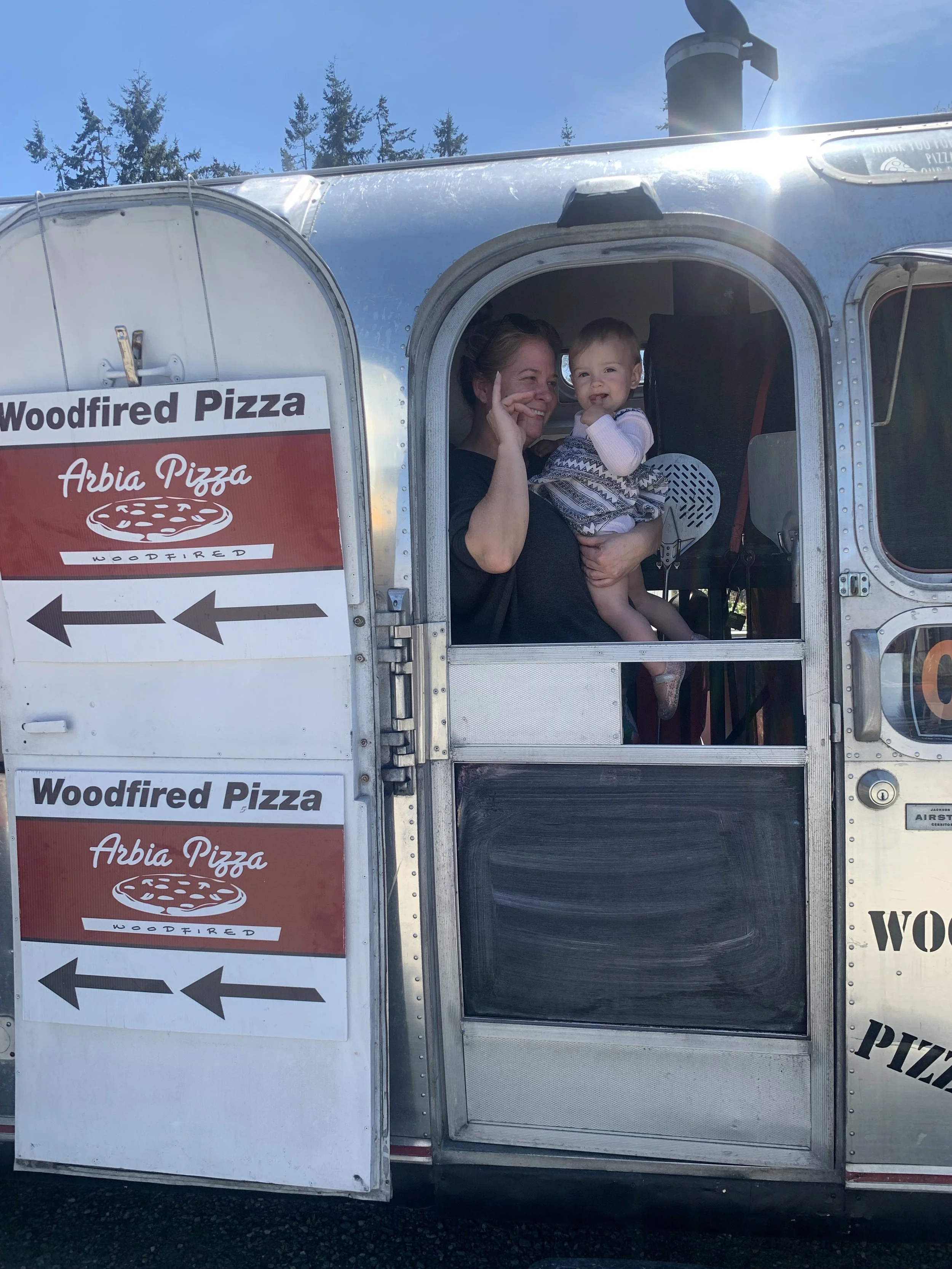 A person holding a child inside a food truck named 'Woodfired Pizza,' with a sign pointing left and trees and blue sky in the background.