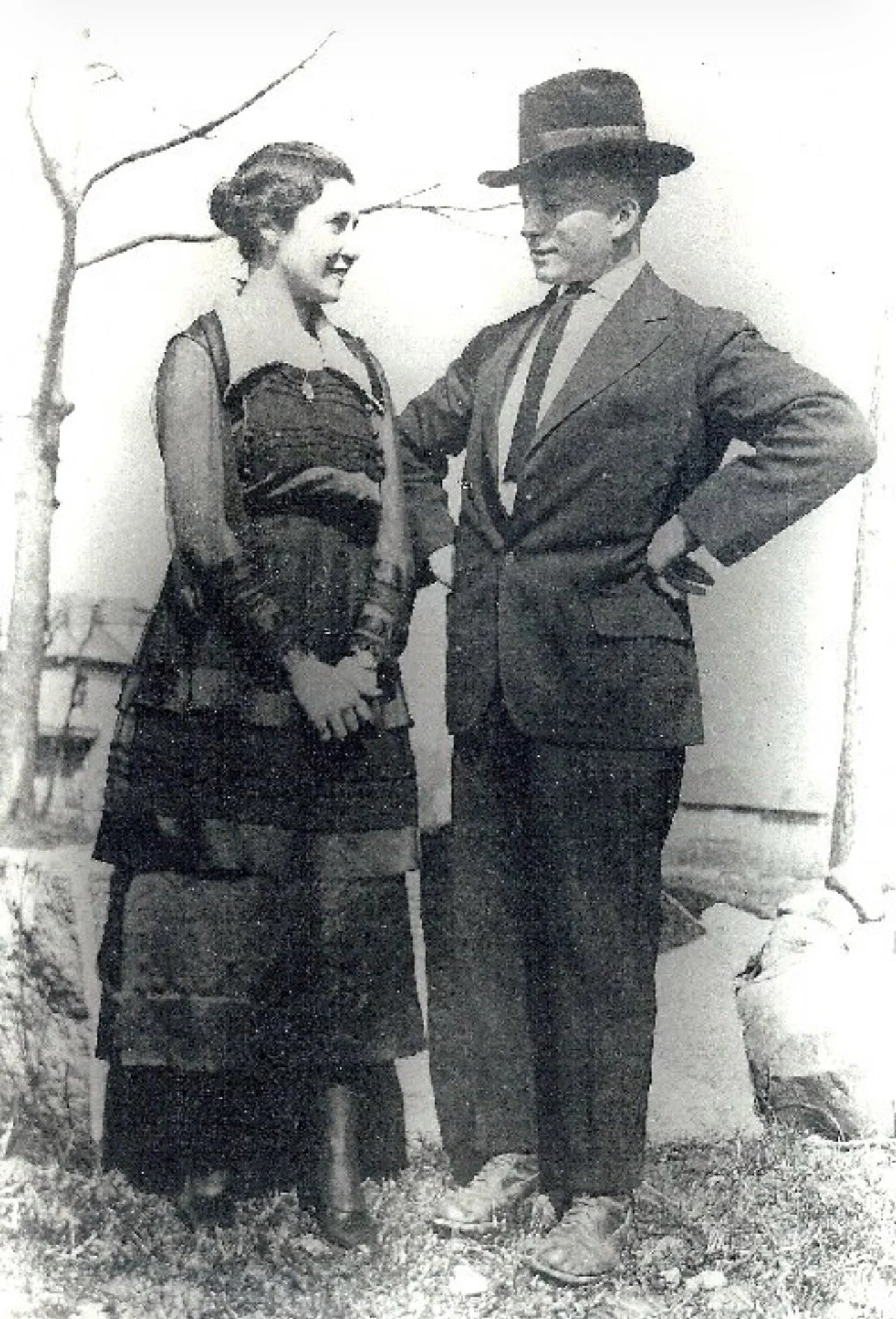 A black and white photo of a man and woman standing on snow, smiling at each other. The woman wears a dress and a jacket, holding an umbrella, and the man wears a suit with a tie and a hat.