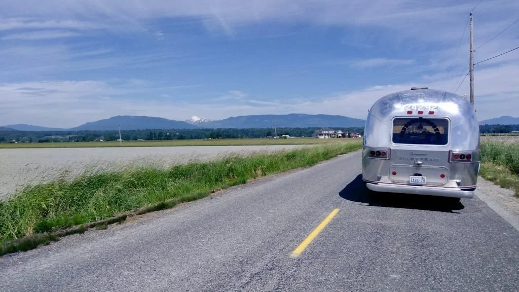 A silver vintage camper trailer on a rural road surrounded by fields with mountains in the distance under a blue sky.