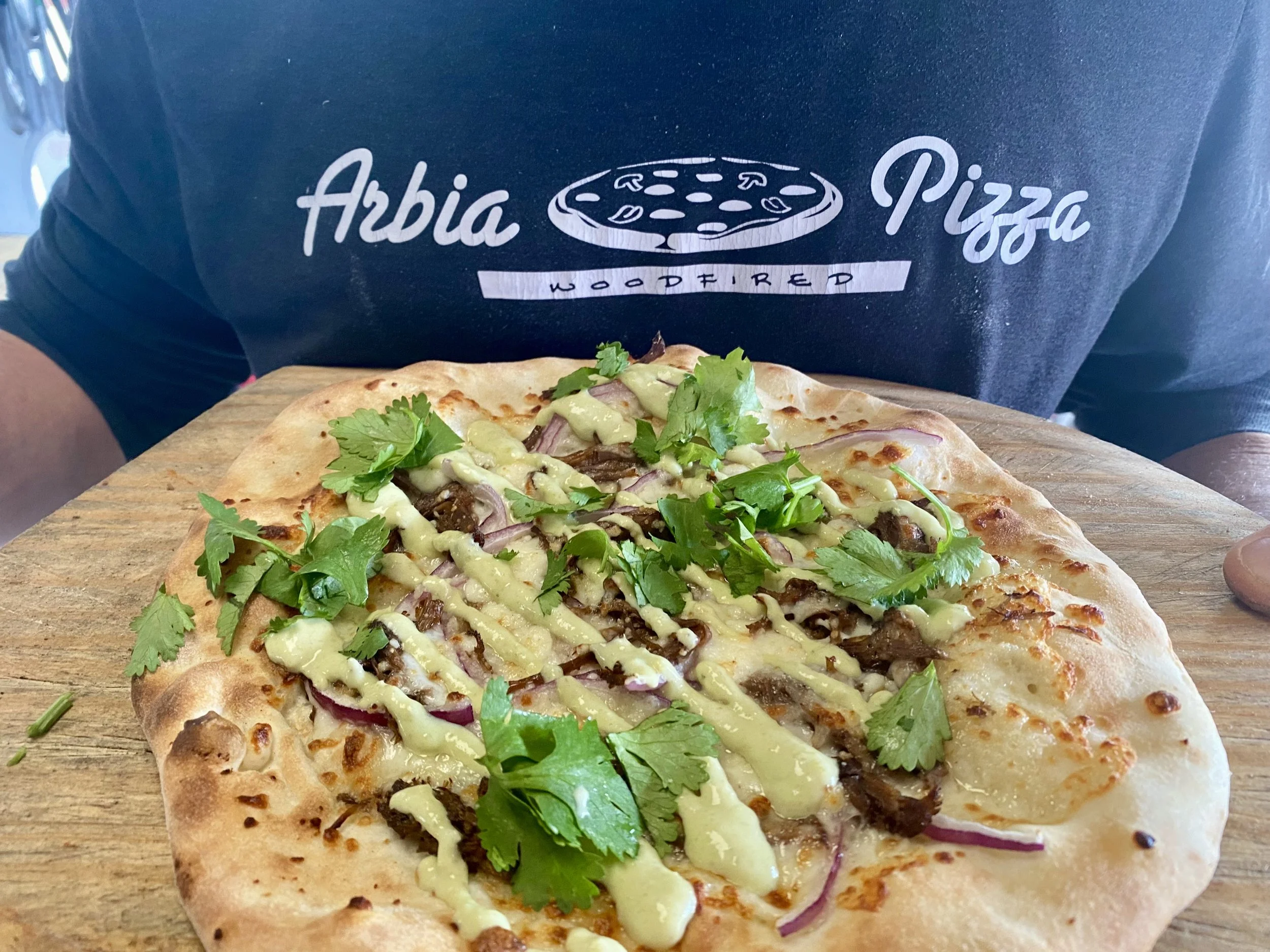A freshly baked pizza with cheese, sliced onions, cilantro, and barbecue beef on a wooden board, with a person wearing a dark shirt that has the words 'Arbia Pizza' and 'Woodfired' and a pizza image printed on it.
