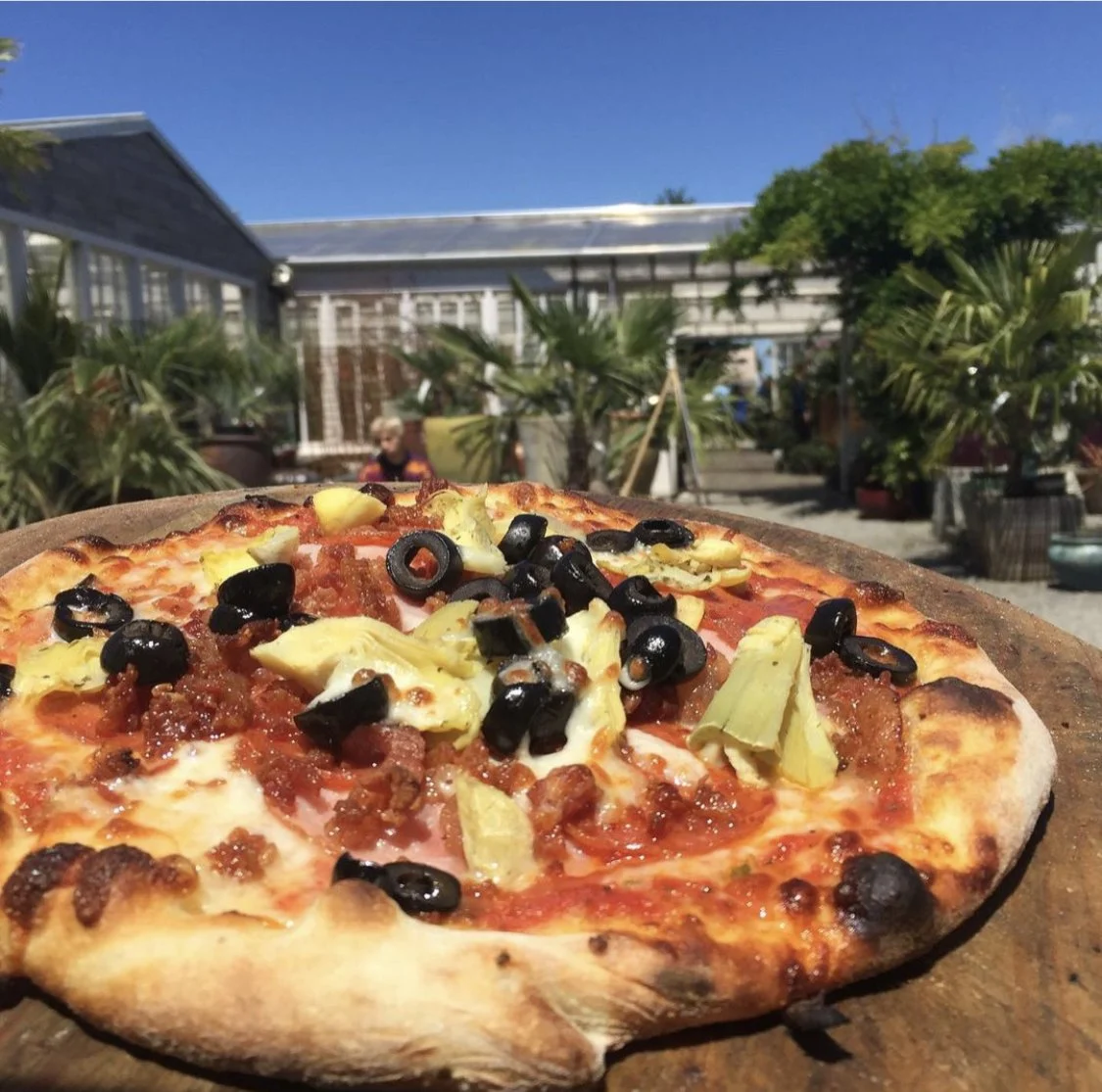 Pizza topped with black olives, artichokes, and tomato sauce on a wooden board outside with a garden and blue sky in the background.