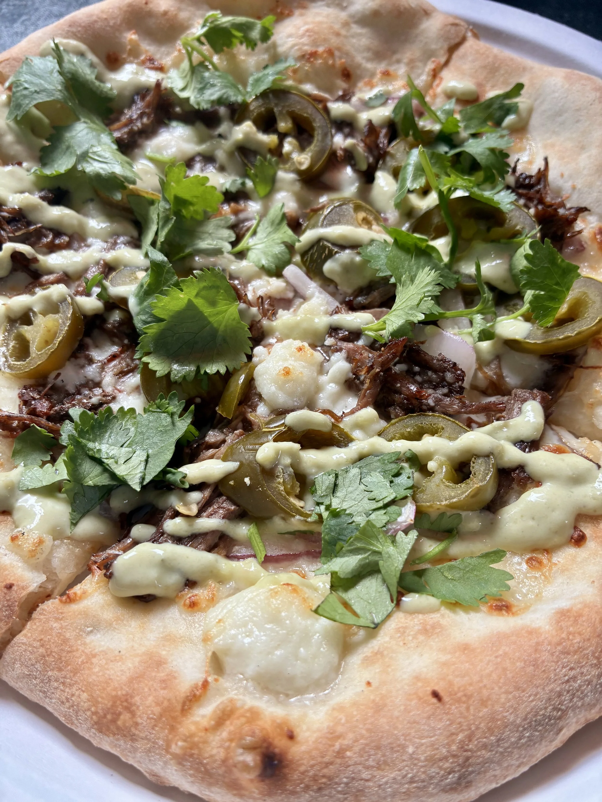 Close-up of a pizza with green herbs, jalapeño slices, shredded meat, white sauce, and melted cheese on a crust.