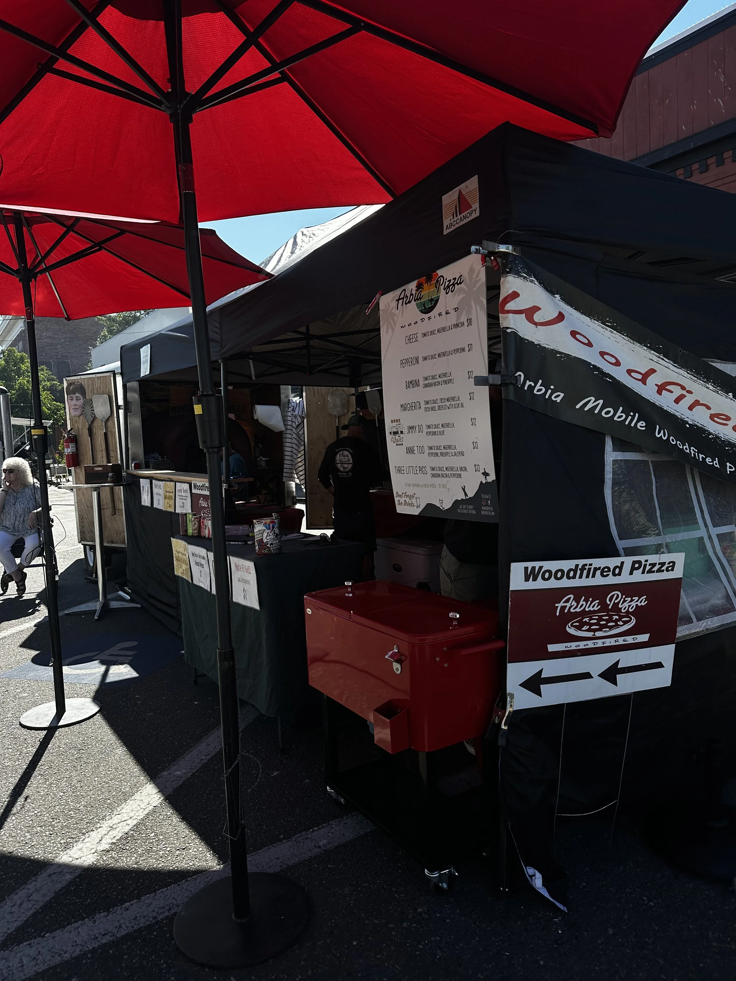 Outdoor food stall with red umbrellas and a menu board for Abia Pizza, Woodfired Pizza, with various pizza options and prices, in a parking lot.