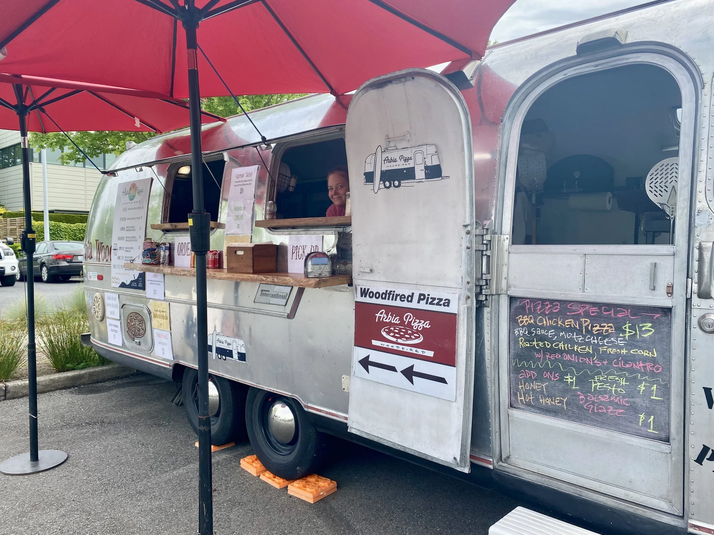 A food truck with a red umbrella and metal exterior, selling pizza, with a woman smiling inside. The truck has chalkboard menus and signs stating 'Woodfired Pizza,' with various pizza options and prices listed.