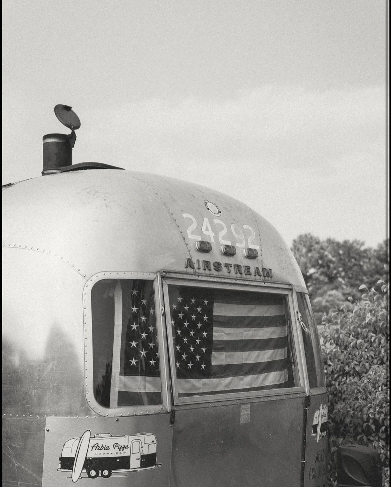 Black and white photo of a vintage Airstream trailer, American flag window, and a sticker for Arbia Pizza