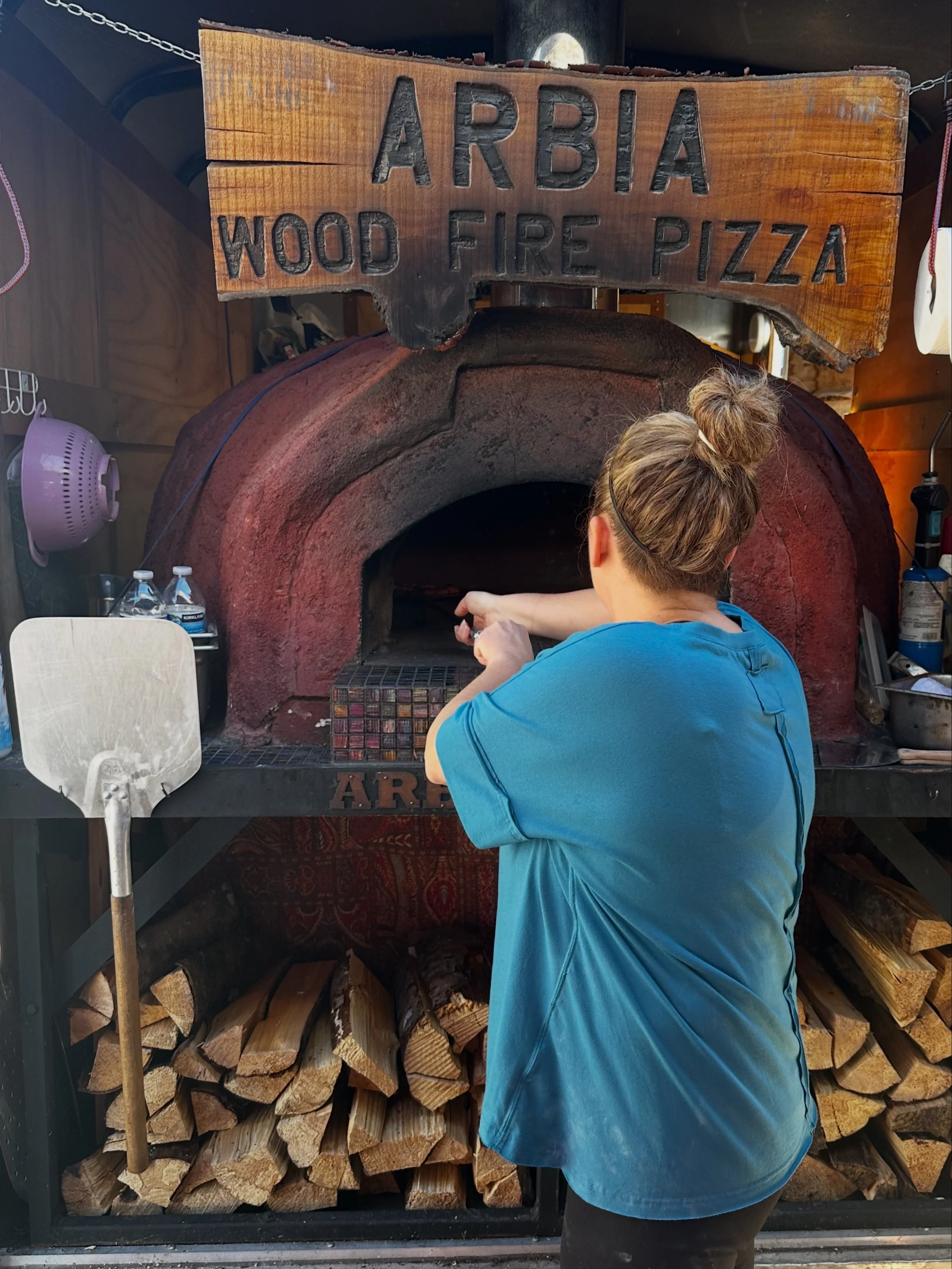 A person in a blue shirt and glasses with a bun in their hair, standing in front of a wood-fired pizza oven. The oven is large and made of brick, with firewood stored underneath. There is a wooden sign above the oven that reads 'Arbia Wood Fire Pizza
