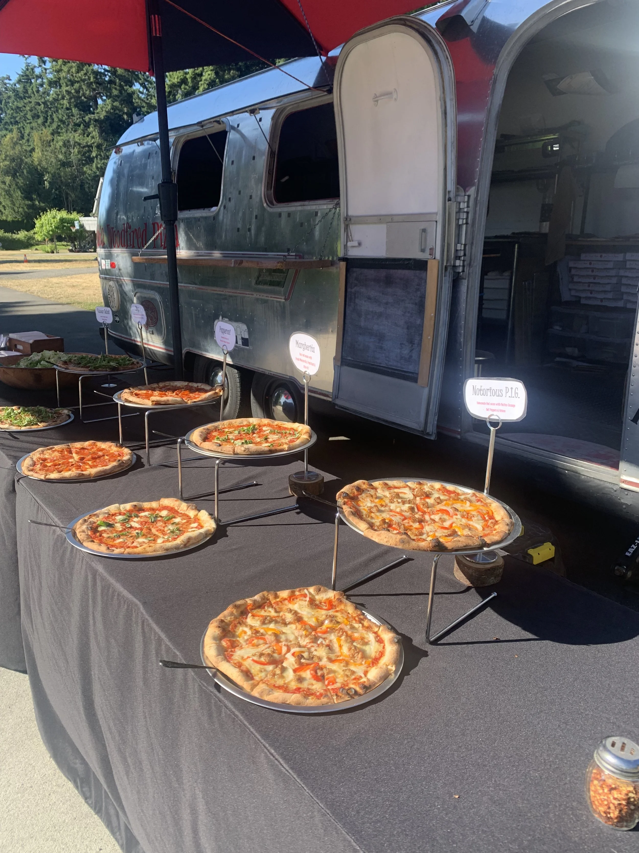 Various pizzas displayed on a table at a food truck event, with a food truck in the background.