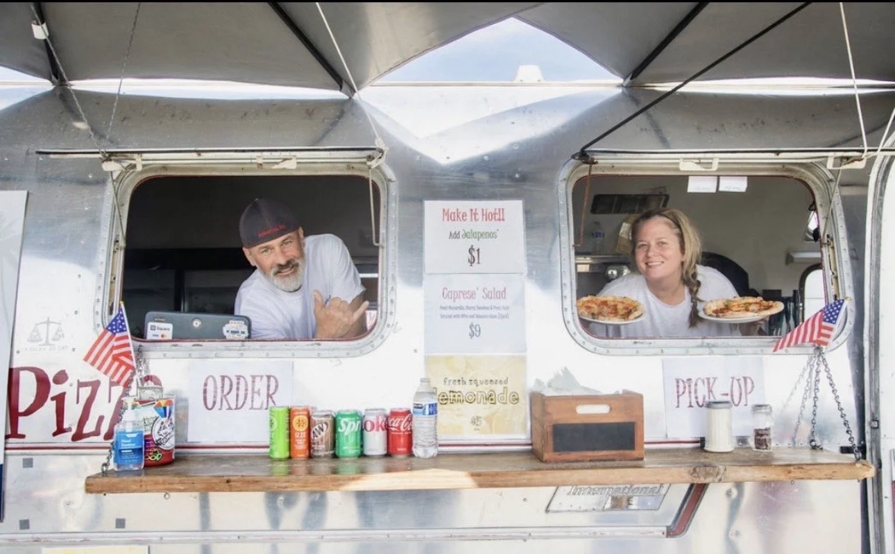 A man and a woman are serving pizza from a food truck. The man is wearing a beanie and white shirt, and is leaning out the window smiling and making a shaka sign. The woman is holding two slices of pizza and smiling. The food truck has signs advertising pizza, salads, and lemonade, with American flags decorated on the window.