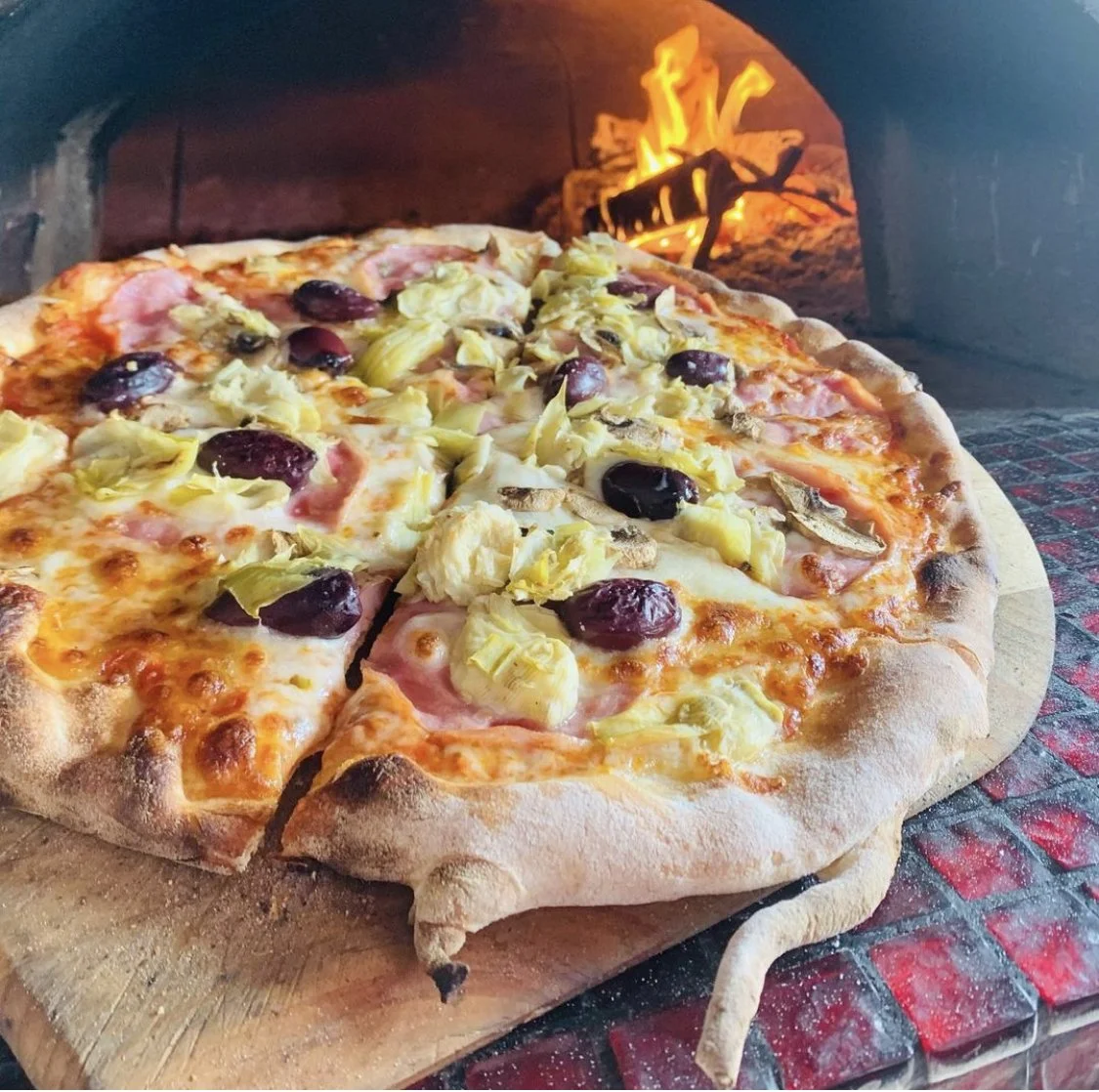Pizza with toppings of black olives, artichokes, mushrooms, ham, and cheese baking in a wood-fired oven.