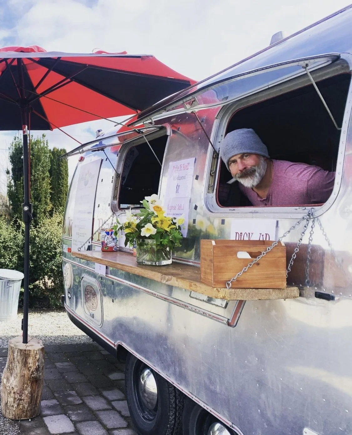 A man with a gray beard and wearing a gray knit cap looks out from a window of a shiny silver food truck. The food truck has a wooden counter with flowers in a glass vase, a small wooden box, and a hanging sign. A large red and black umbrella shades part of the truck. The truck is parked on a paved area with some greenery in the background.