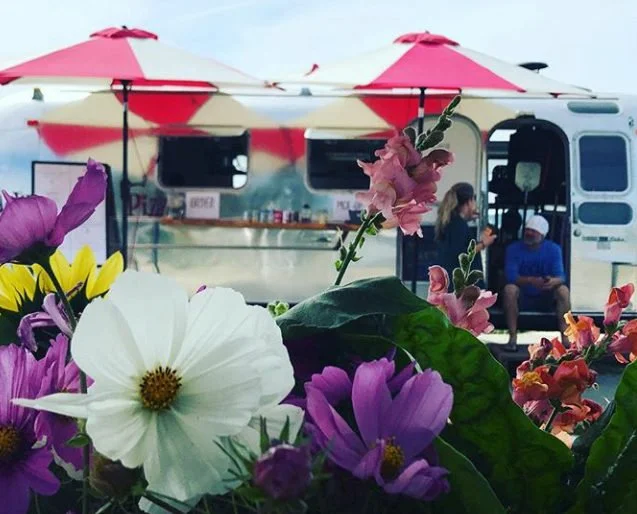 Colorful flowers in front of a food truck with red and white umbrellas, people sitting outside the truck.
