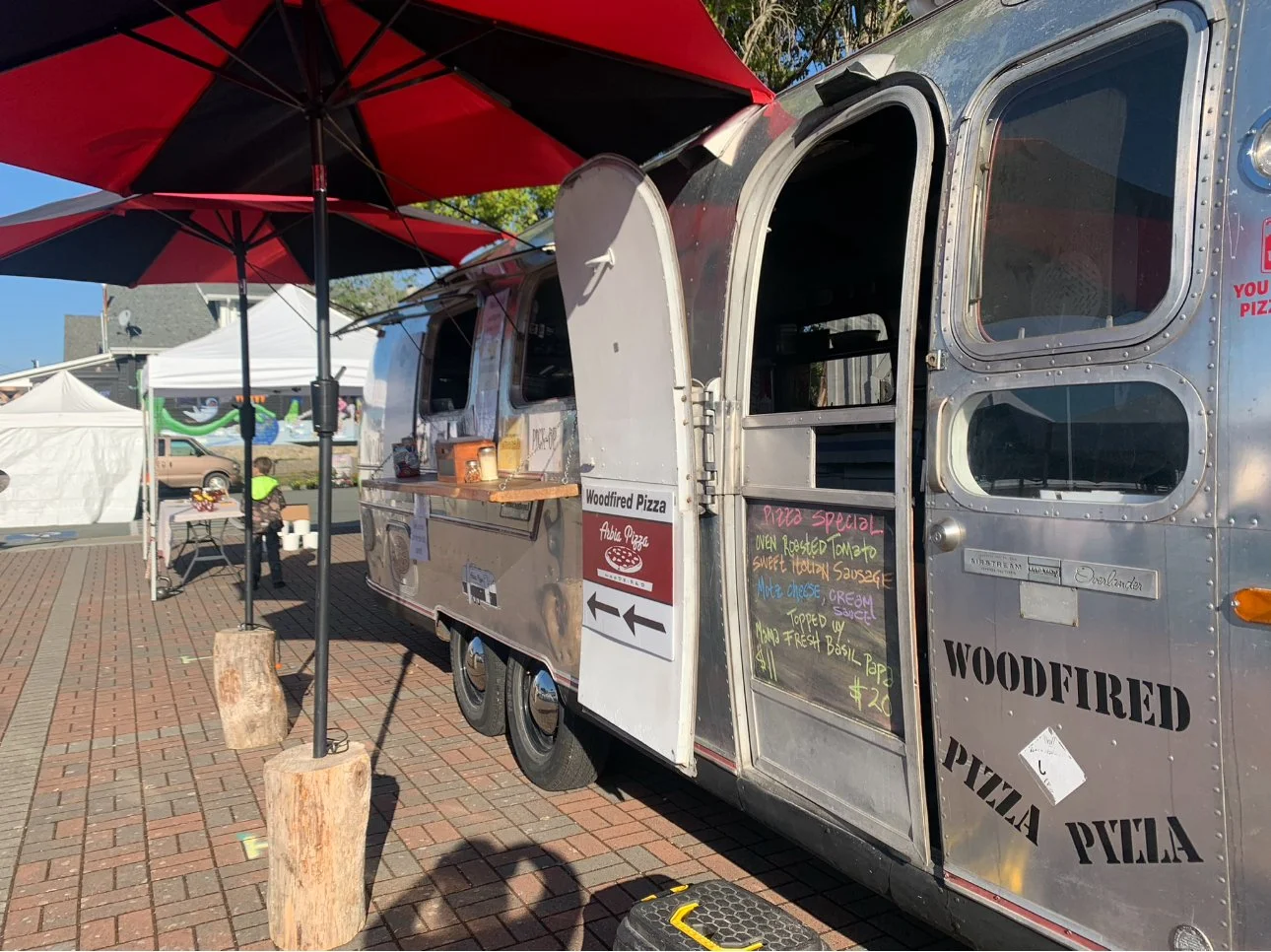 A food truck selling pizza at an outdoor event, with a red and black umbrella, a chalkboard menu, and a tent in the background.