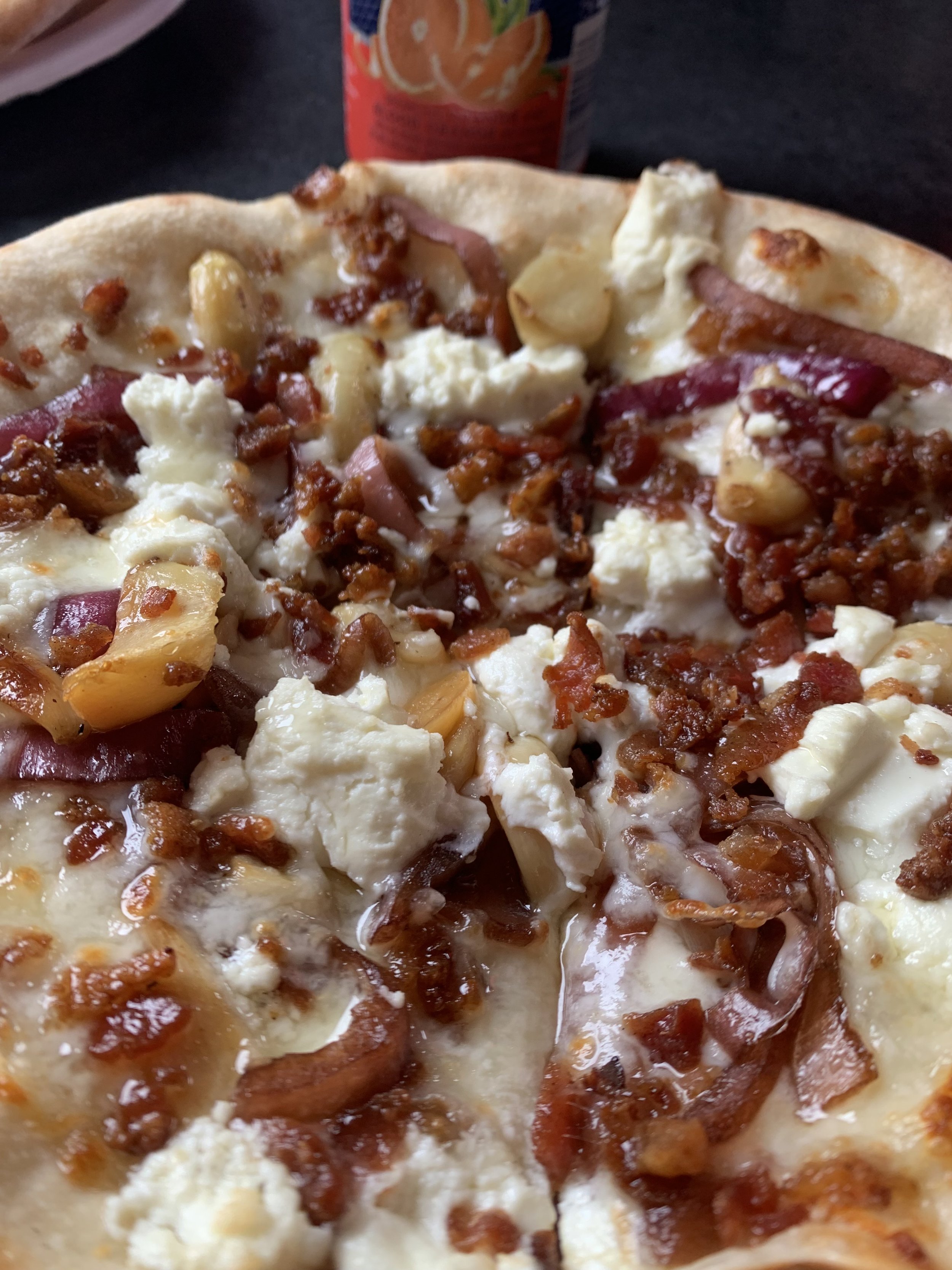 Close-up of a pizza with cheese, sausage, ham, and mushrooms, with a can of orange juice in the background.