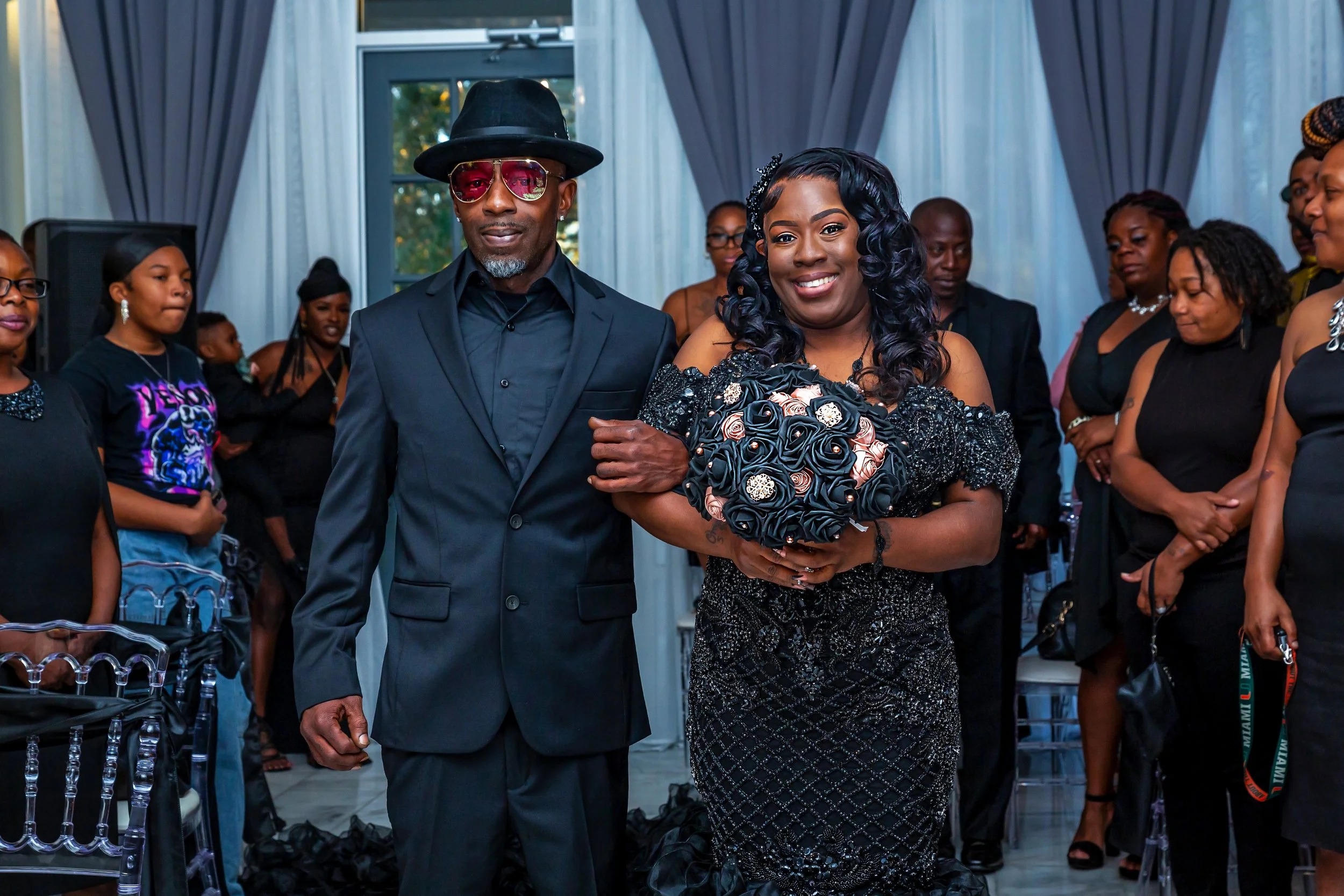 A woman dressed in a black beaded gown holding a bouquet of roses walks down the aisle with an older man in a black suit, who is wearing a black hat and pink sunglasses, during a wedding ceremony in a decorated indoor space with group of guests watch