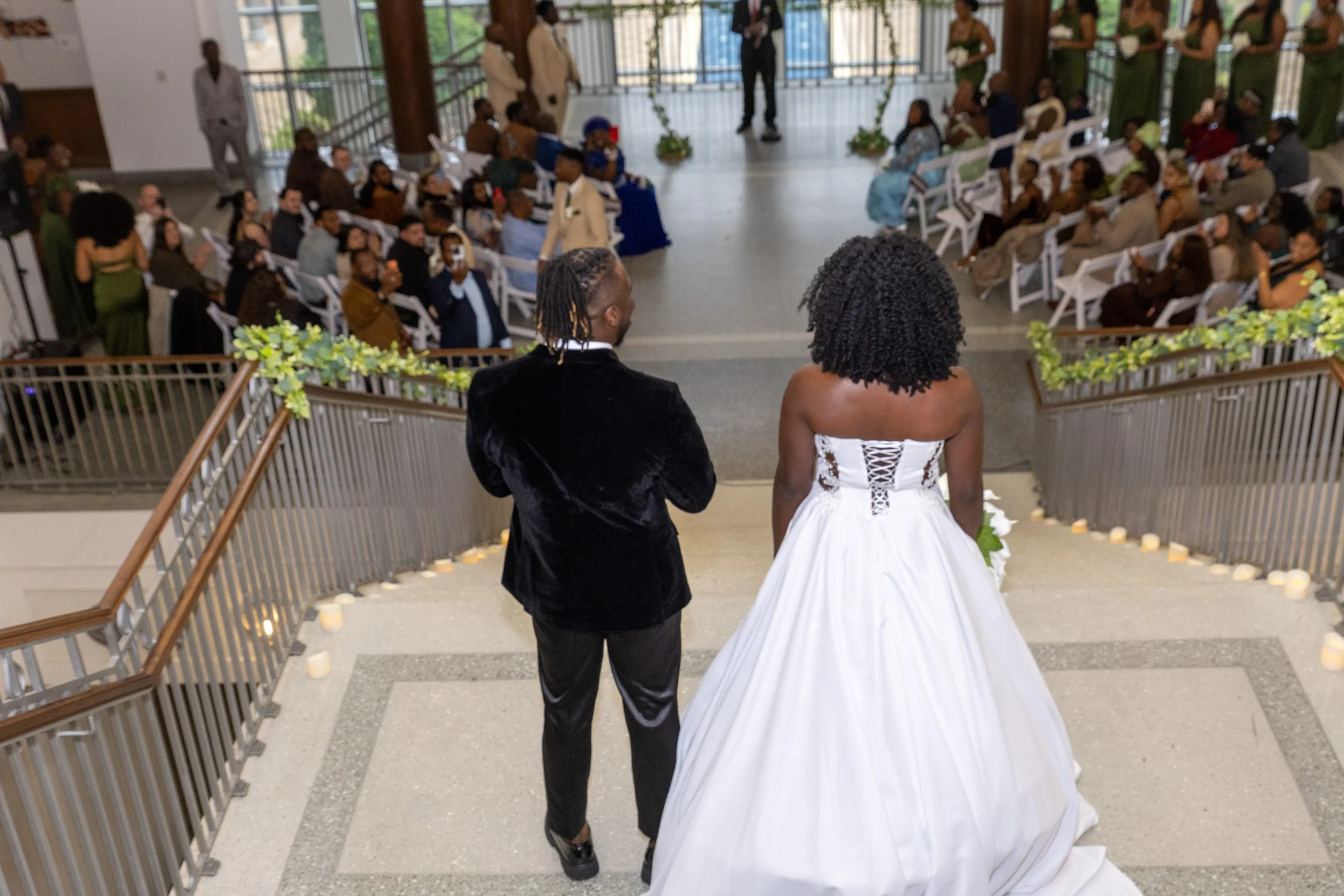 A couple, the man in a black tuxedo and the woman in a white wedding gown, stands at the top of a staircase, overlooking guests seated for a wedding ceremony. The scene is indoors with natural light coming through large windows, and the hallway is de