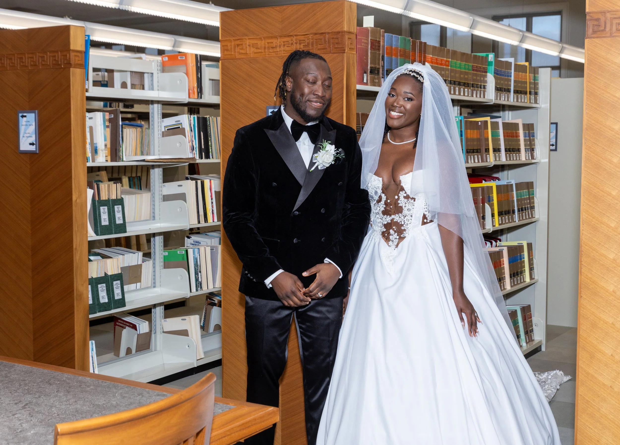 A bride and groom standing together in a library surrounded by bookshelves, with the bride in a white wedding dress and veil, smiling, and the groom in a black tuxedo with a boutonnière, holding hands.