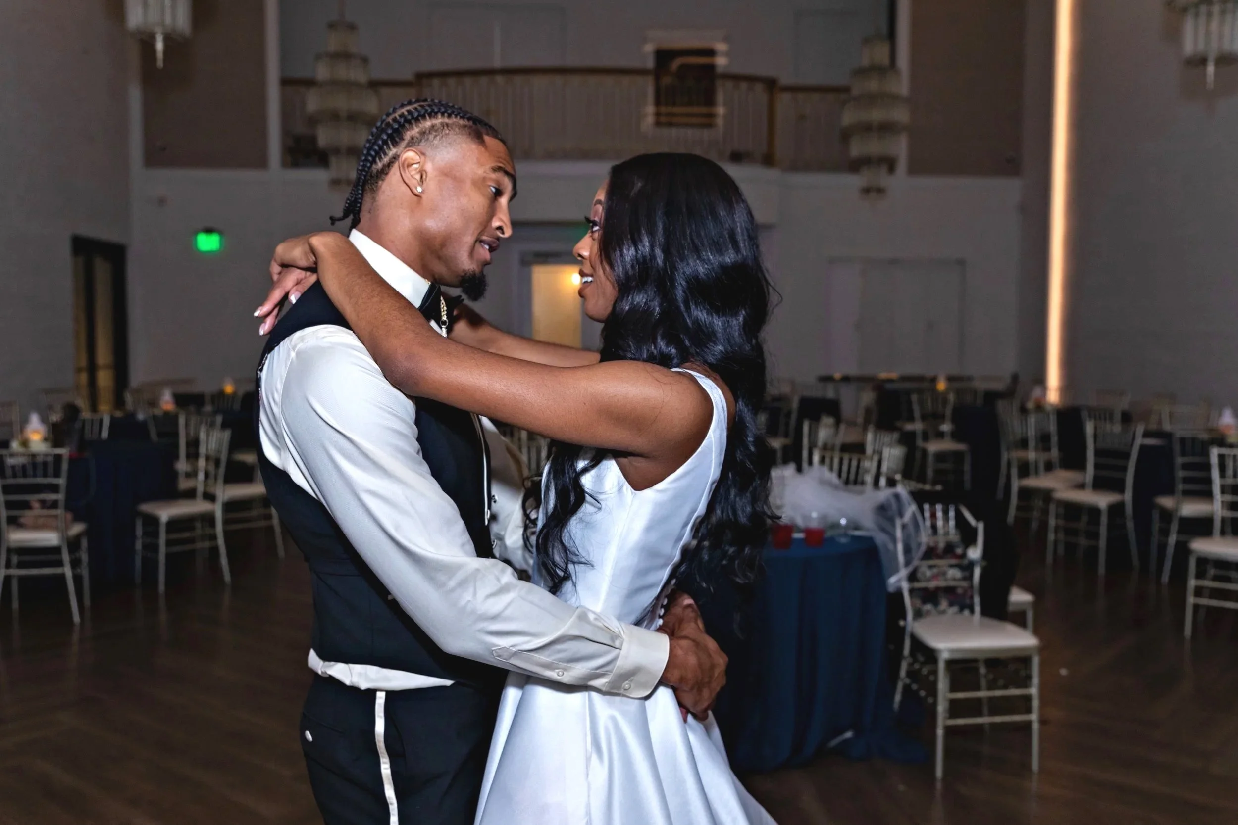 A young couple dancing closely at a formal event in a decorated banquet hall with tables and chairs.