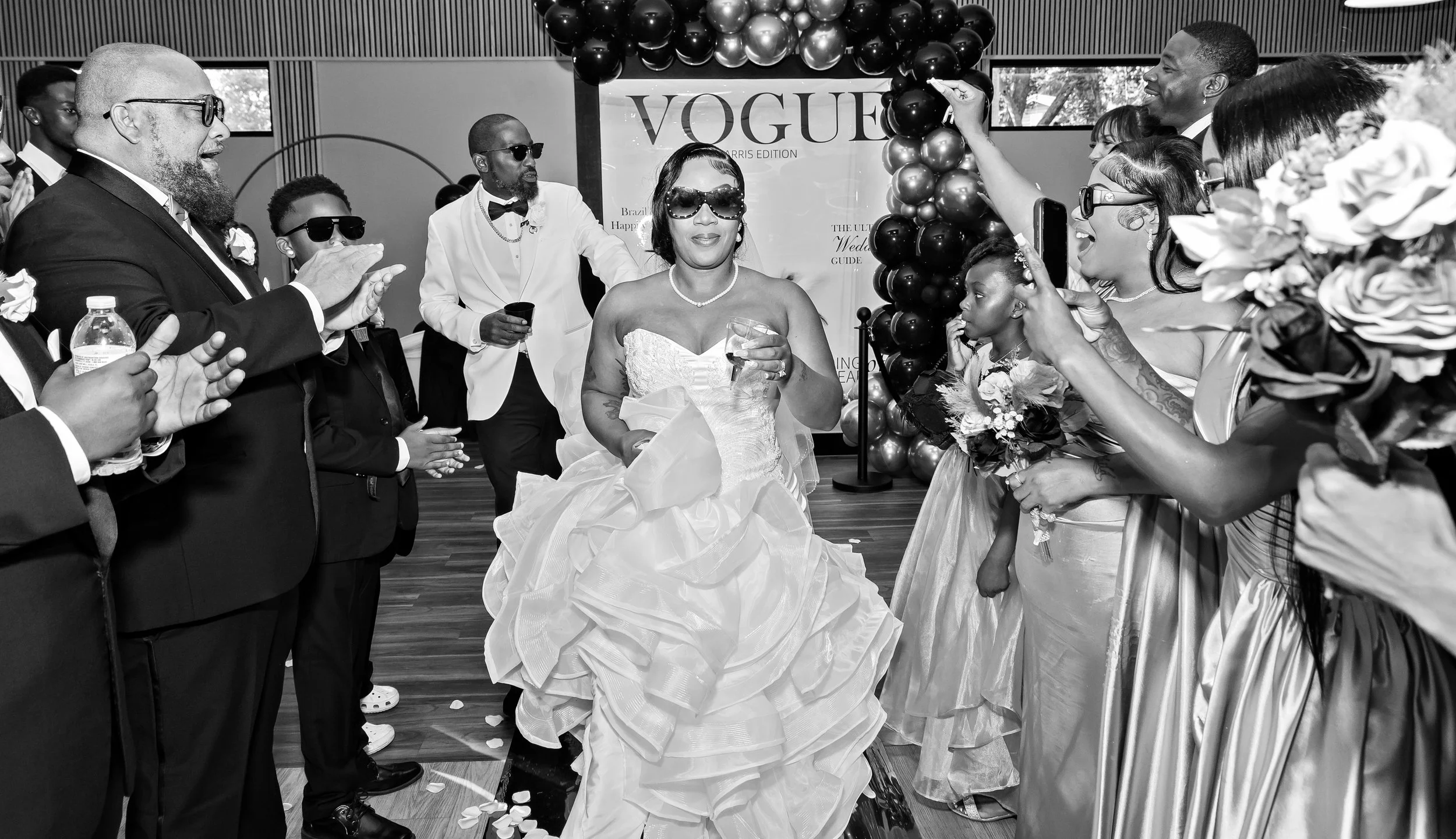 A group of people at a celebration, with a woman in a wedding dress in the center, wearing sunglasses, holding a glass, surrounded by family and friends clapping and smiling. There is a Vogue sign with balloons in the background.