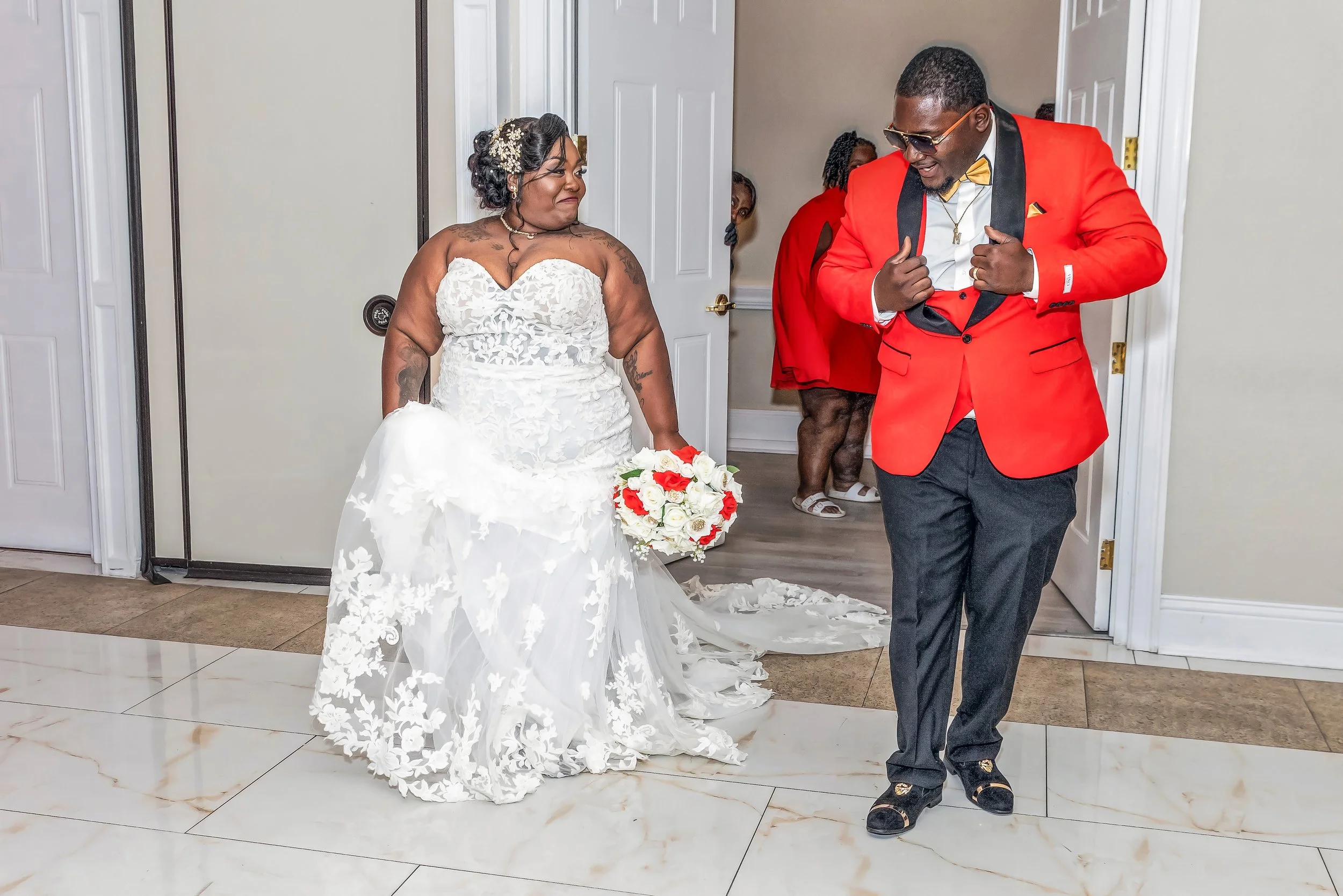 A bride in a white strapless wedding gown holding a bouquet of white and red roses, standing next to a man in a red suit jacket with a black lapel, black pants, and sunglasses, as they share a joyful moment during a wedding celebration indoors.