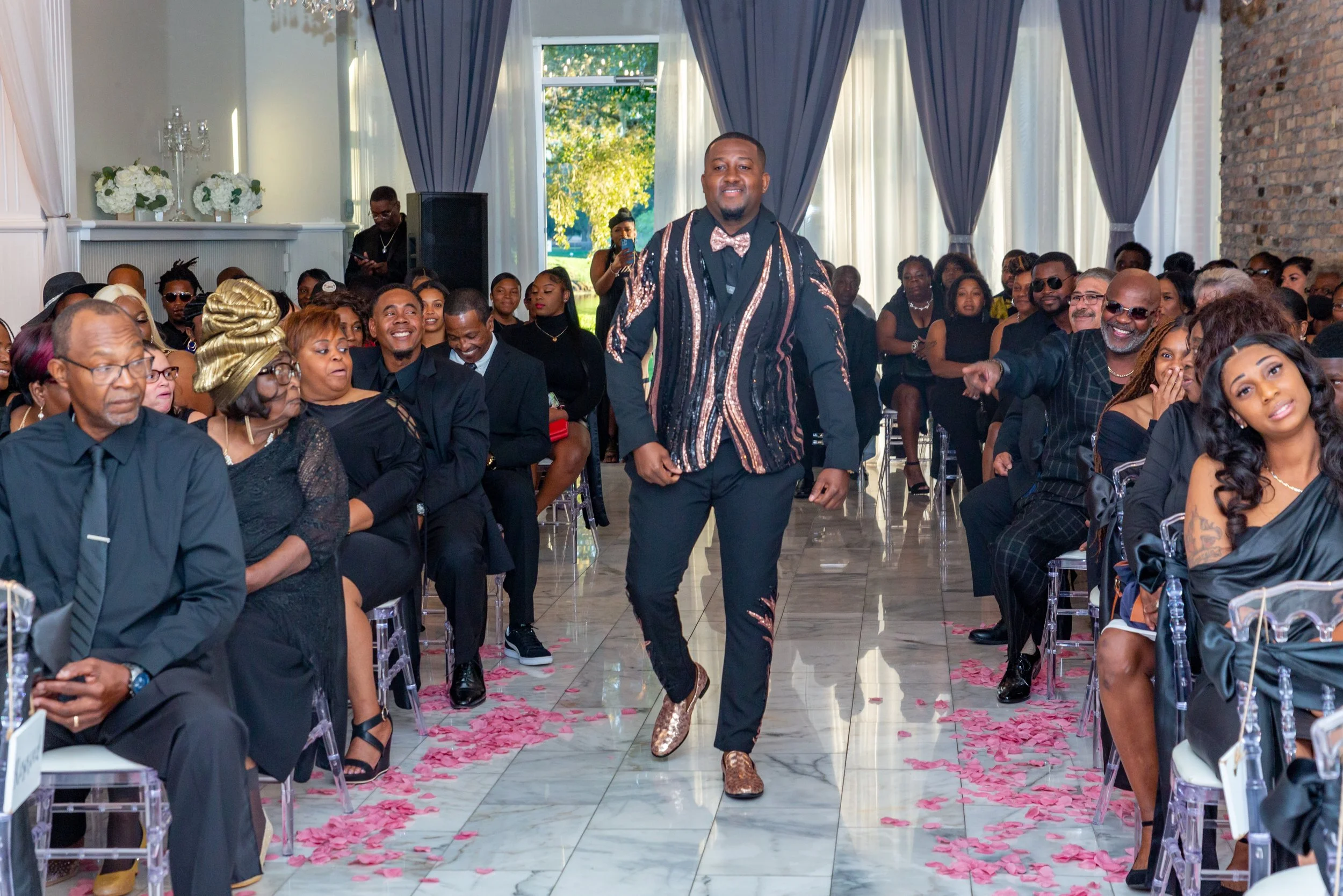 Man walking down aisle in a formal event, surrounded by seated guests, some smiling and some with neutral expressions, pink flower petals scattered on the floor, decorated with curtains and floral arrangements.