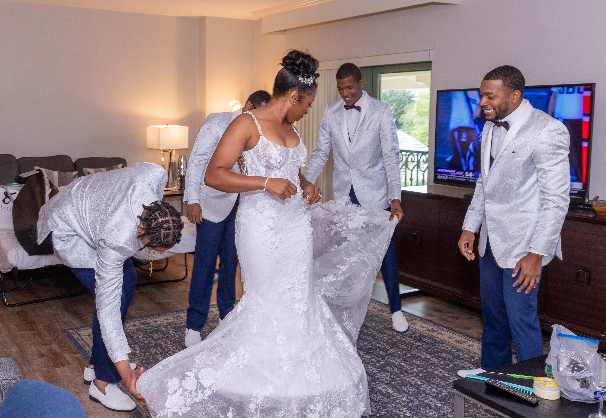 A wedding dress inside a room being adjusted by several people, with a television and balcony in the background.