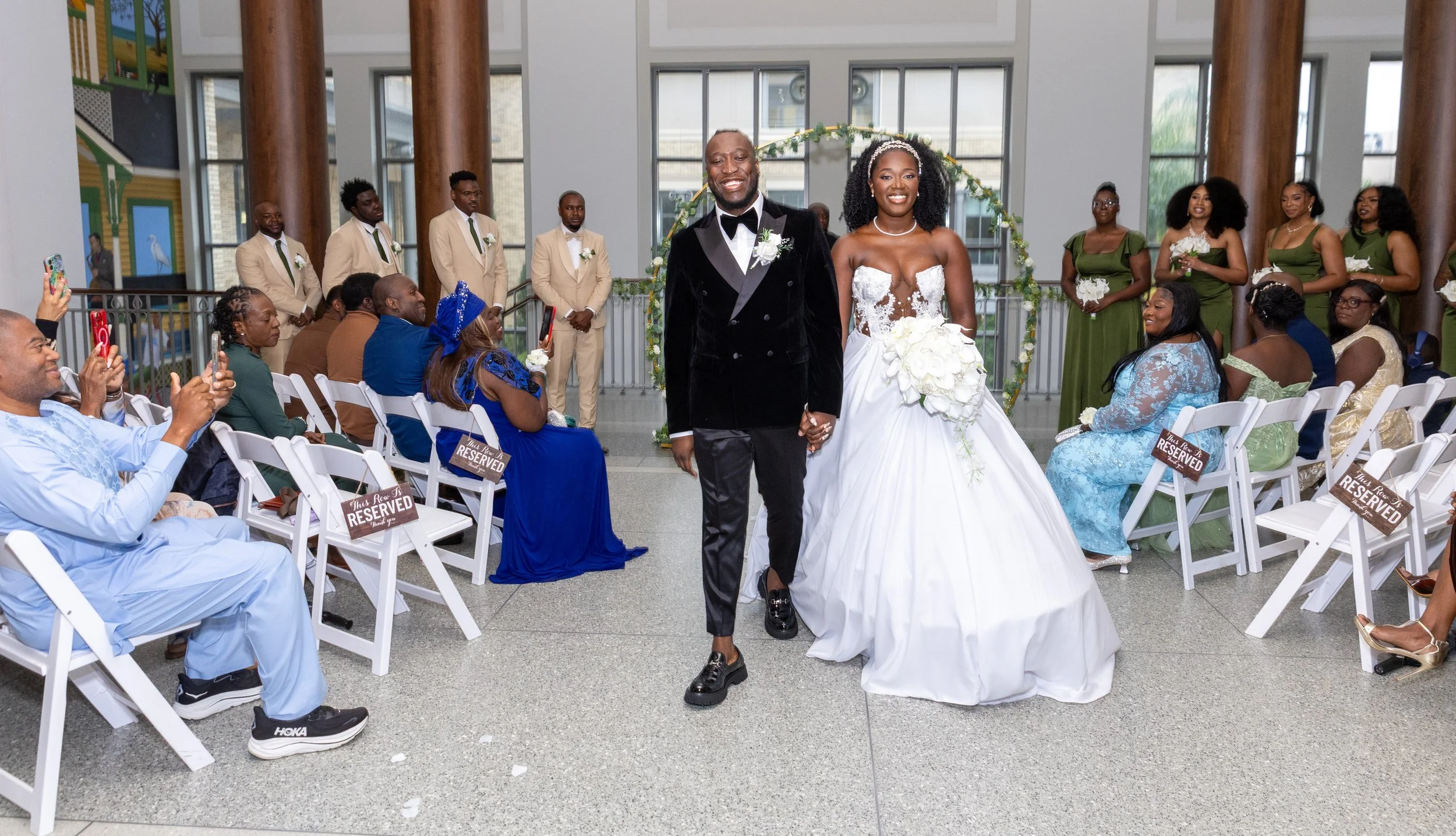 A wedding ceremony with a bride and groom walking hand in hand down the aisle, surrounded by seated guests and bridesmaids in green dresses. The indoor venue has large windows, wooden columns, and a floral arch in the background.