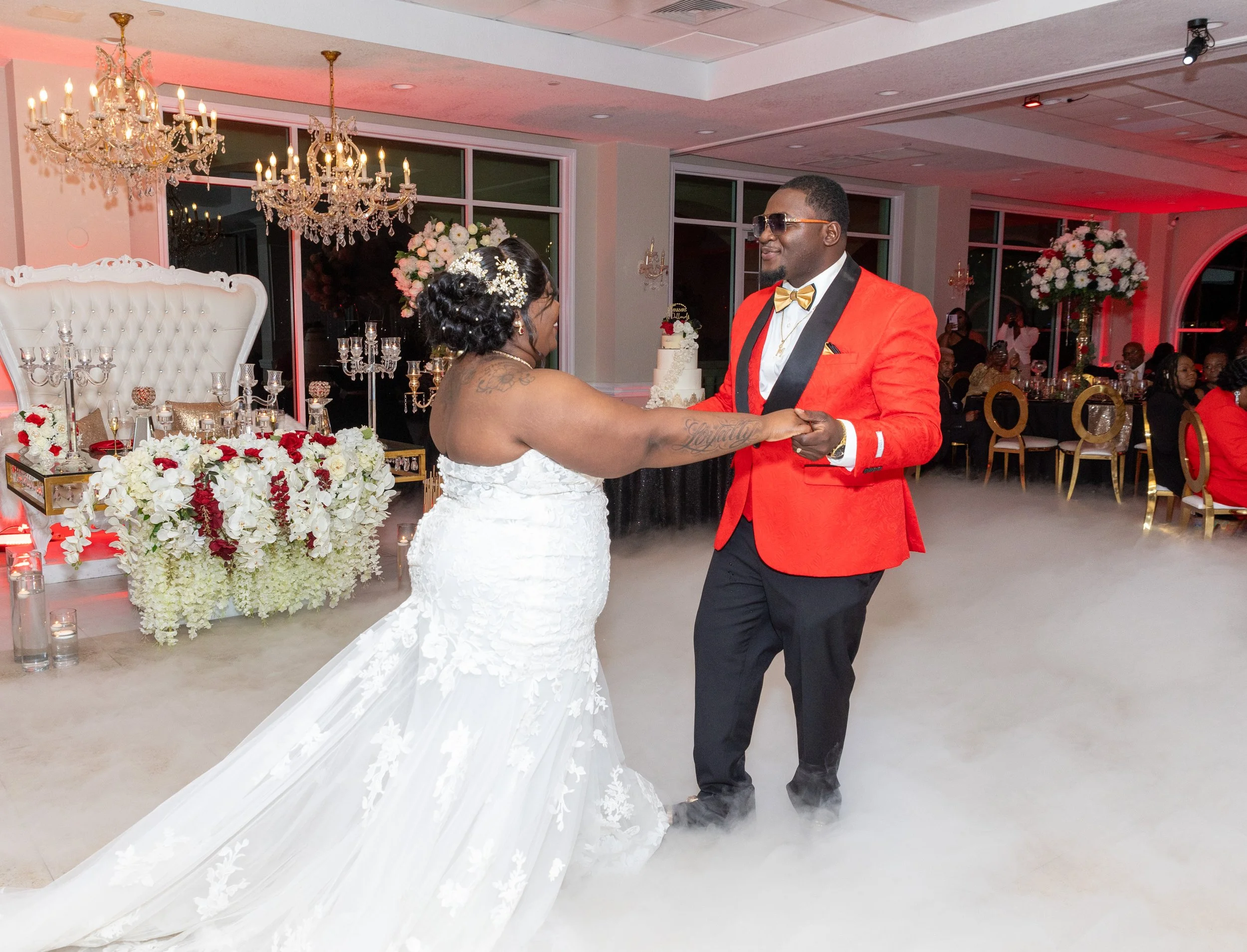 A bride and groom dancing at their wedding reception. The bride is wearing a white wedding dress with floral appliqué, and the groom is dressed in a red tuxedo jacket with a gold bow tie. The background features elegant chandeliers, floral arrangemen