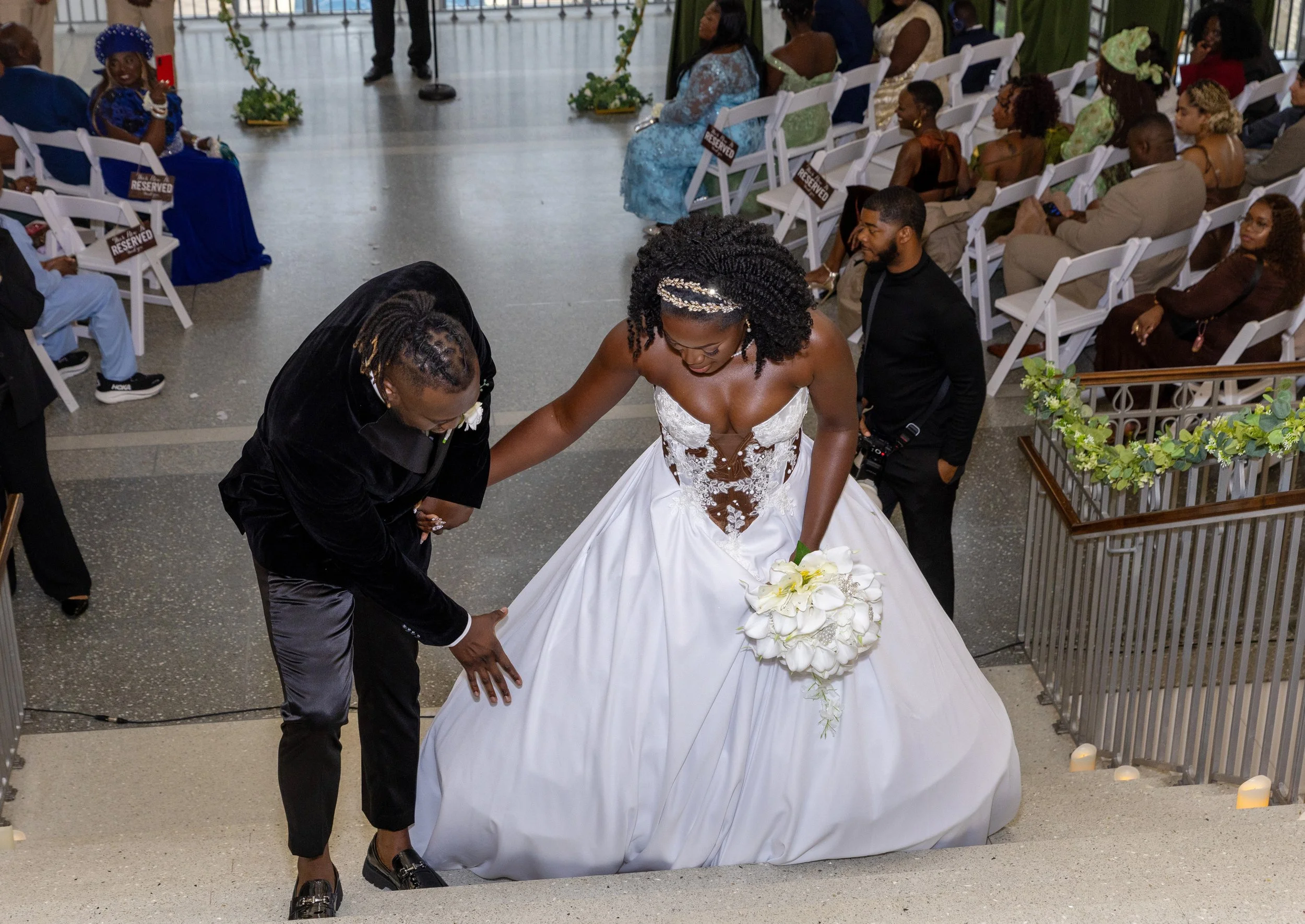 A bride in a white wedding dress with lace details is walking down the aisle with a man in a black tuxedo, holding her hand and looking down at her. Guests seated on white chairs are watching, with some holding phones. The venue is decorated with flo