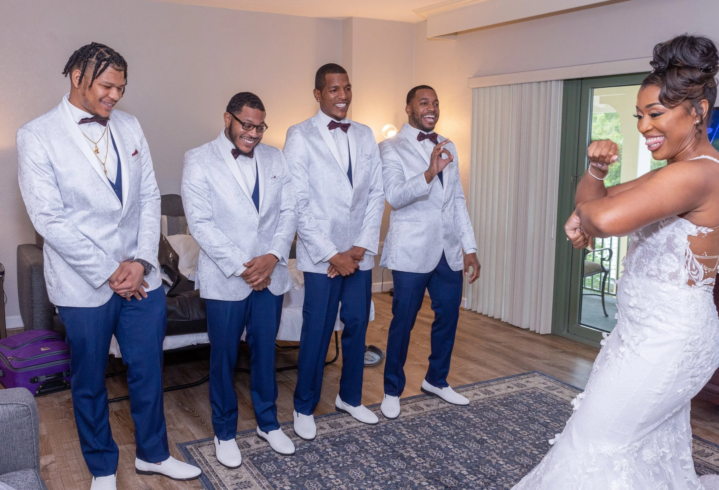 A bride showing her arm and smiling at five groom candidates dressed in matching tuxedos with white jackets, blue pants, purple bow ties, and white shoes, during a wedding preparation.
