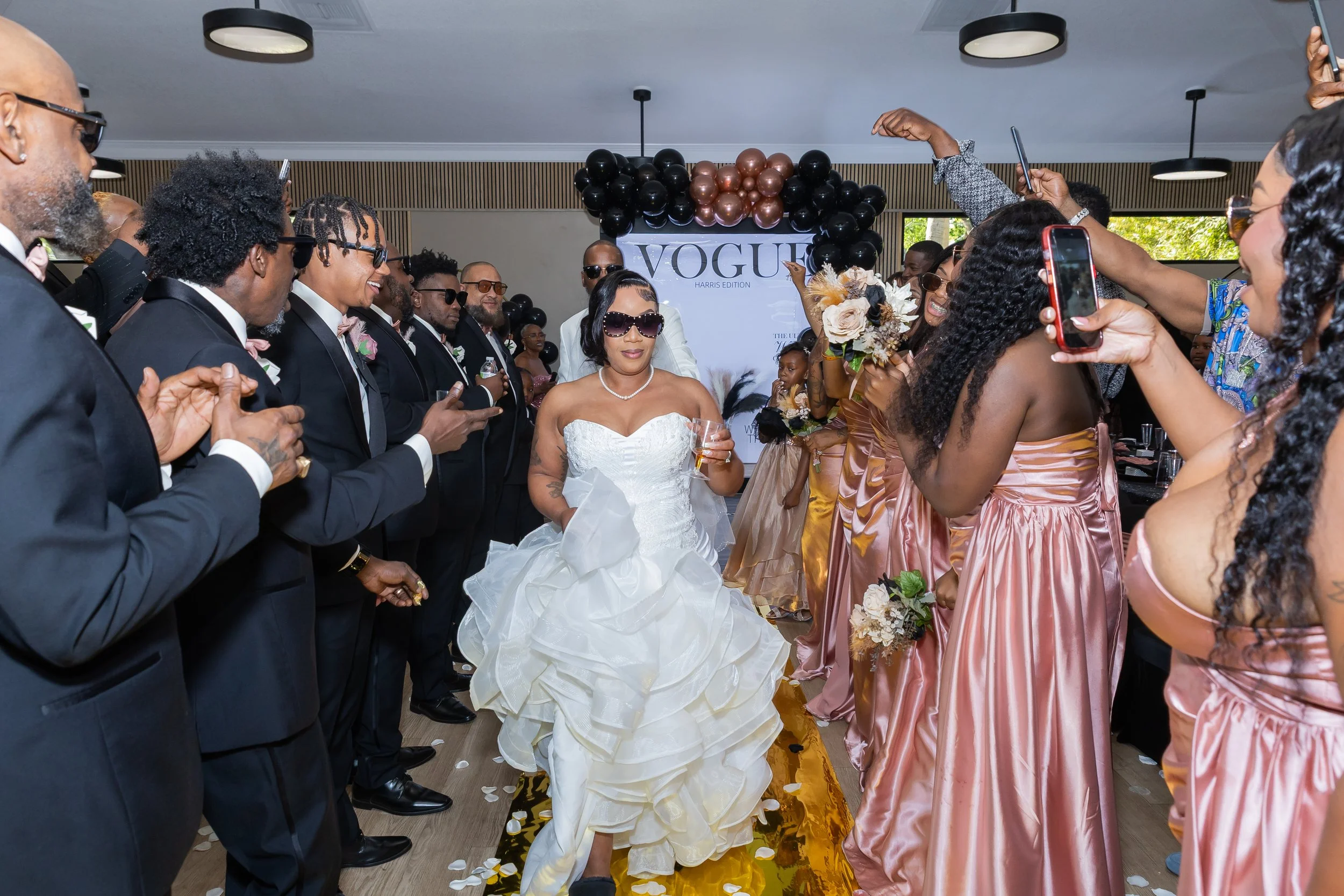 Bride in a white wedding dress holding a glass, surrounded by people in formal attire at a wedding reception with black and metallic balloons and a Vogue sign in the background.