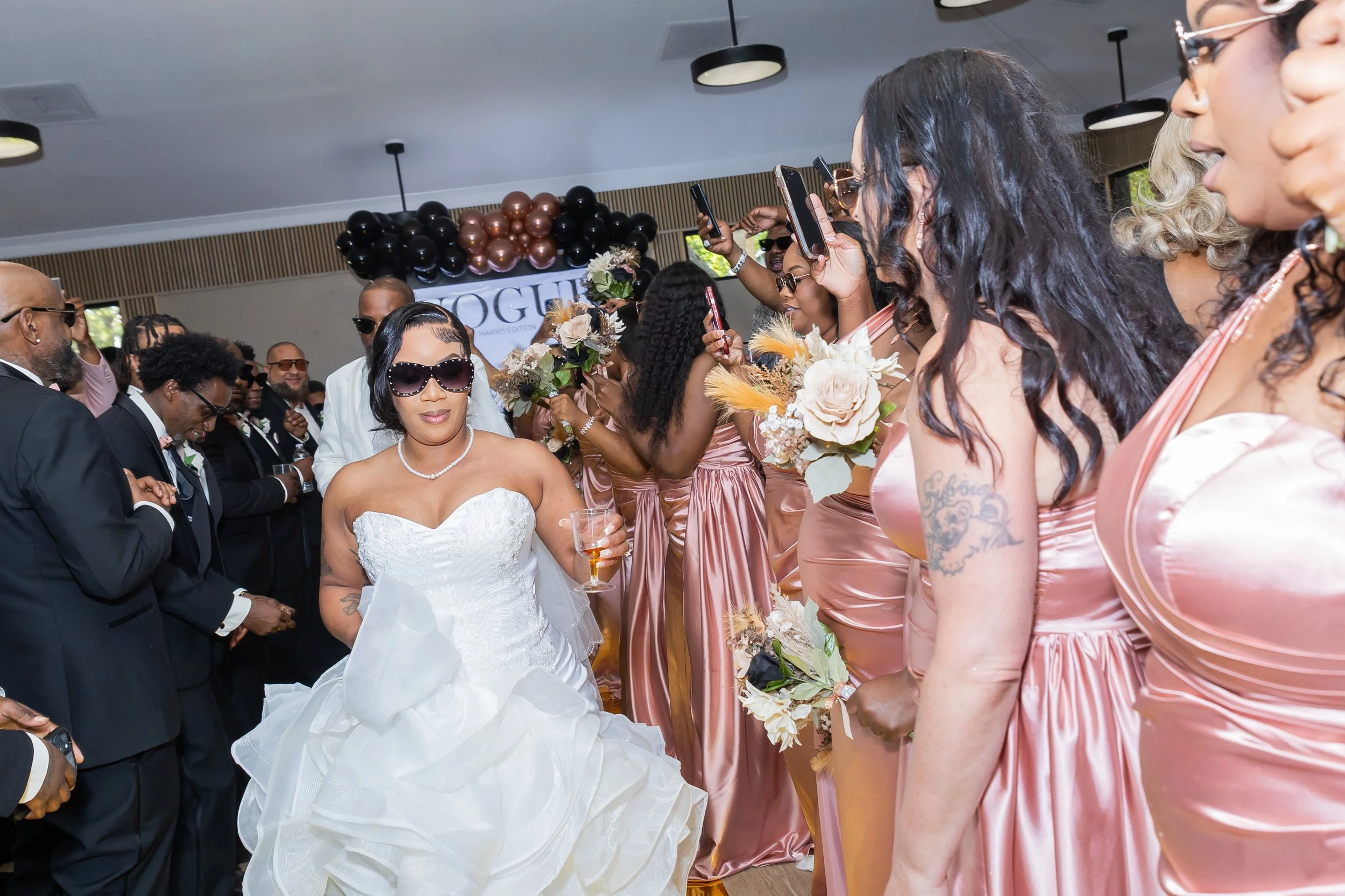 A bride in a white wedding dress holding a glass, surrounded by women in matching pink satin dresses holding bouquets, at a wedding reception with a black and metallic balloon decoration and a Vogue sign in the background.