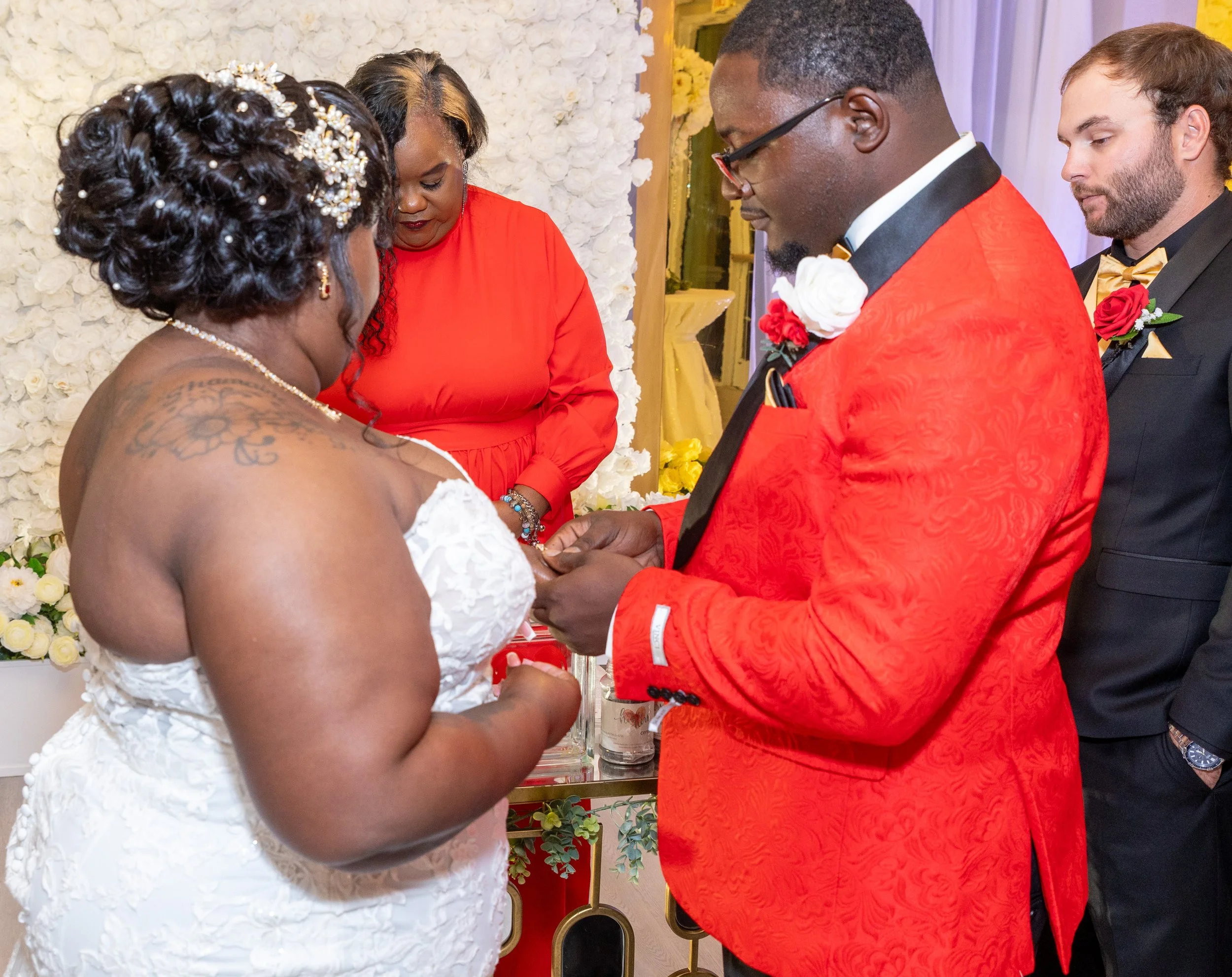 A wedding ceremony with a bride and groom exchanging rings, with an officiant and groomsman present, in front of a white floral backdrop.