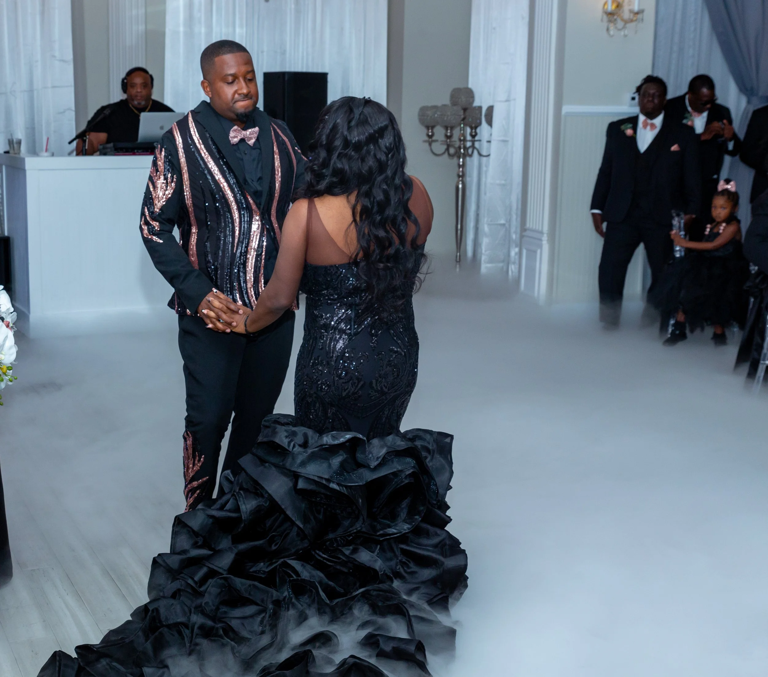 A man and woman are dancing together at a formal event, with a man DJing in the background. The woman is wearing a long black, sparkly gown with a ruffled train, and the man is dressed in a black tuxedo with a sequined jacket and bow tie. Other guest