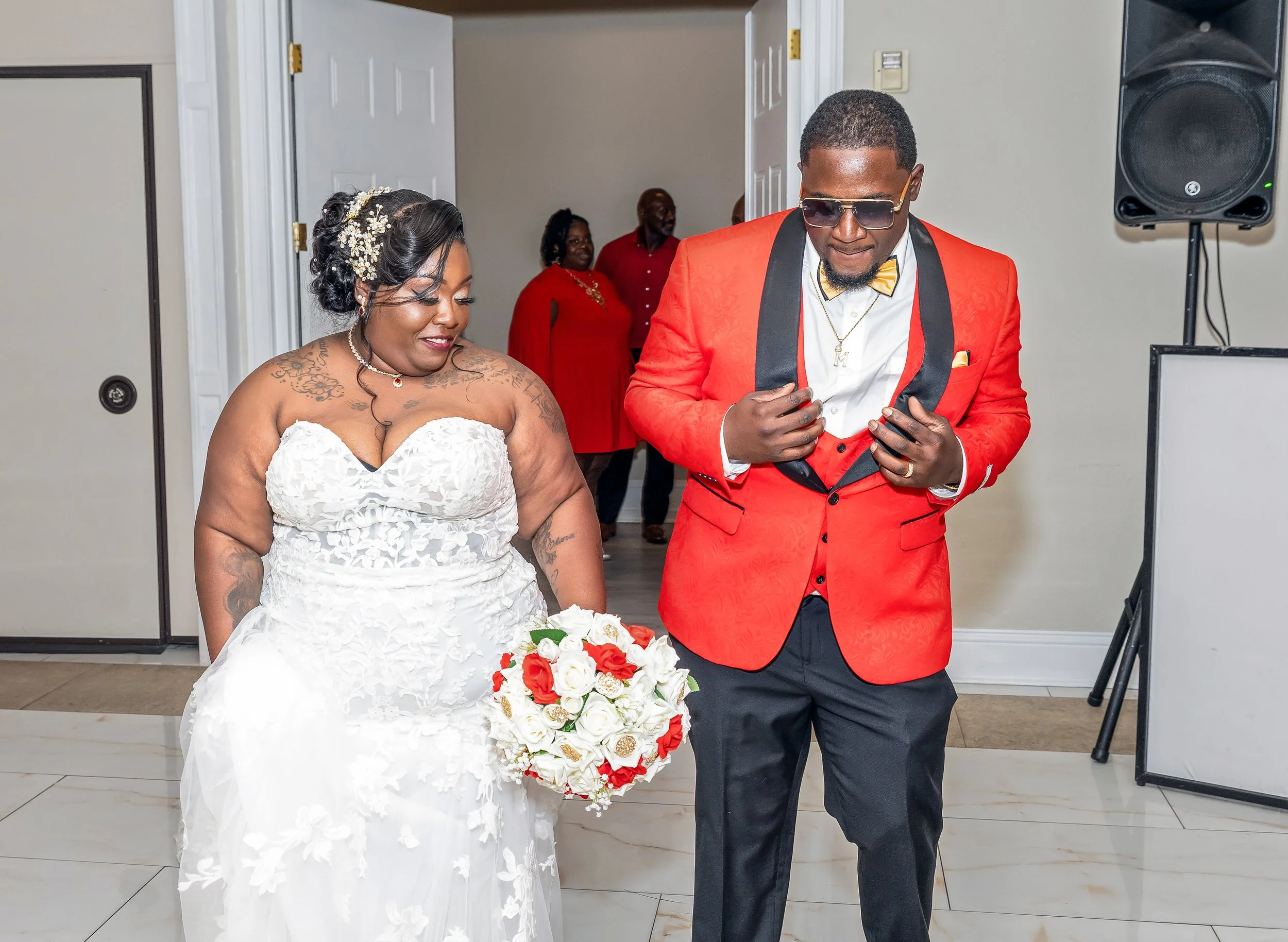 A bride and groom at a wedding ceremony; the bride is wearing a white lace wedding dress, floral hair accessories, and has several tattoos on her arm, holding a bouquet of white and orange flowers. The groom is dressed in a bright red tuxedo jacket, 