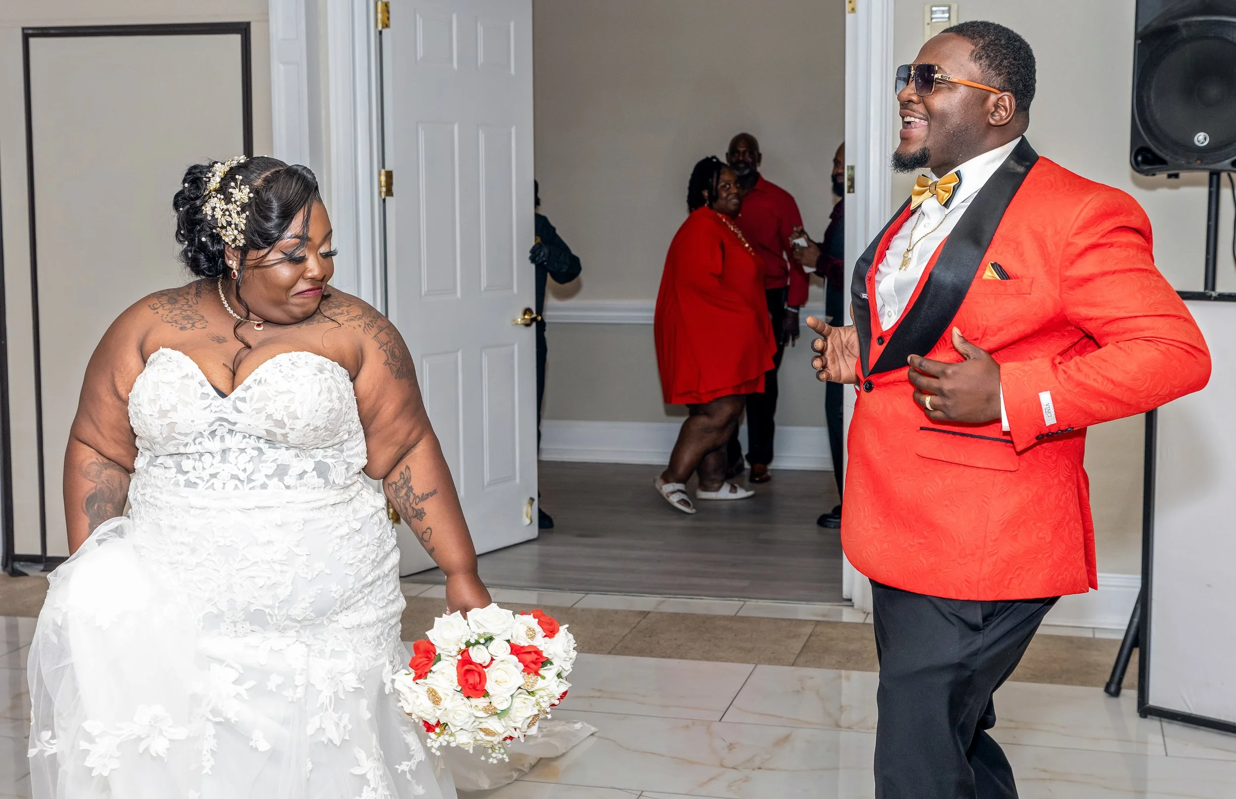 A bride in a white wedding dress with tattoos on her arms, holding a bouquet of white and red roses, standing across from a smiling man in a red tuxedo jacket with black lapels, black pants, and sunglasses, at a wedding ceremony indoors, with guests 