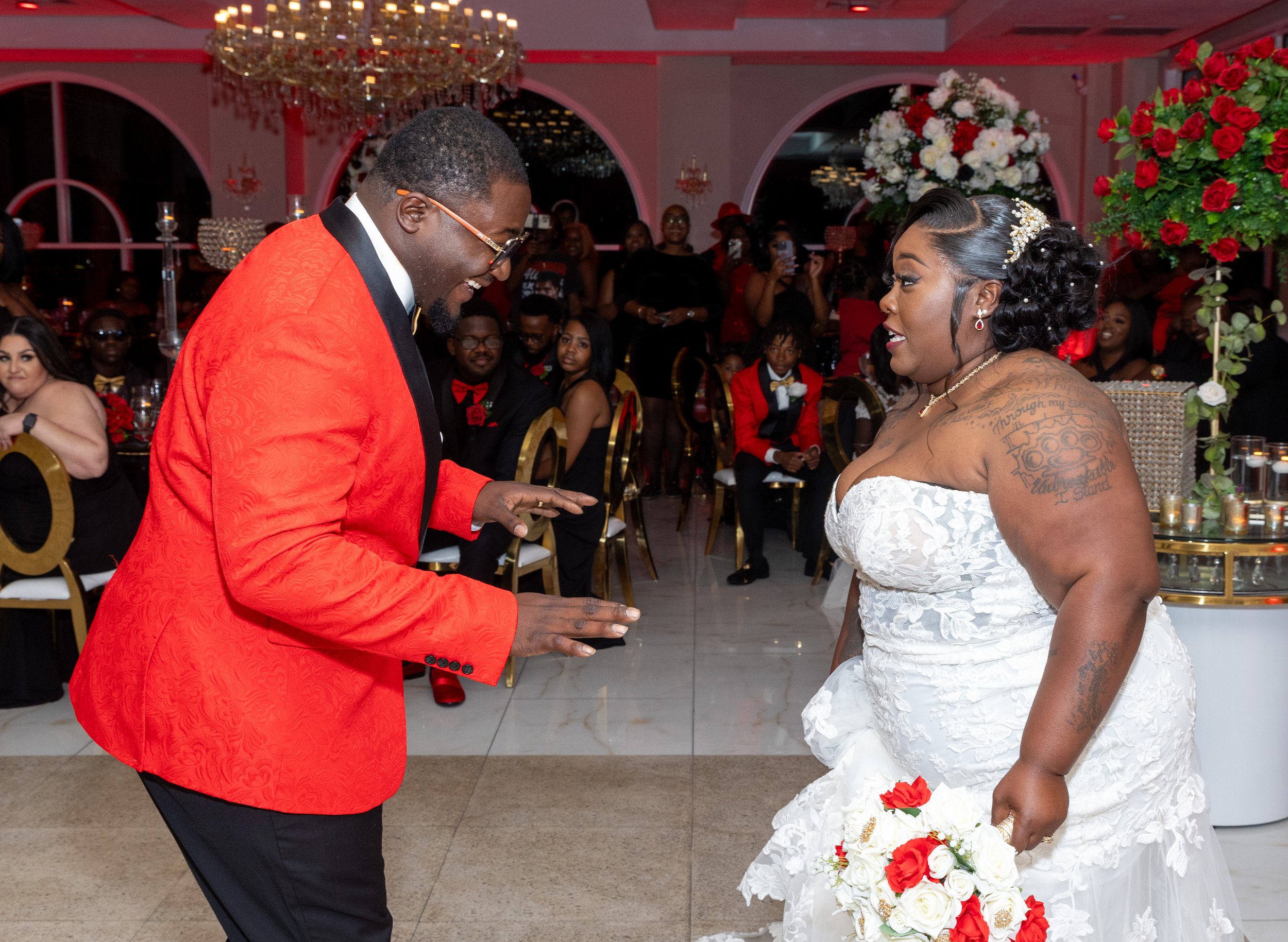 A groom in a red suit is exchanging rings with a bride in a white wedding dress during their wedding ceremony, surrounded by guests in a decorated indoor venue.