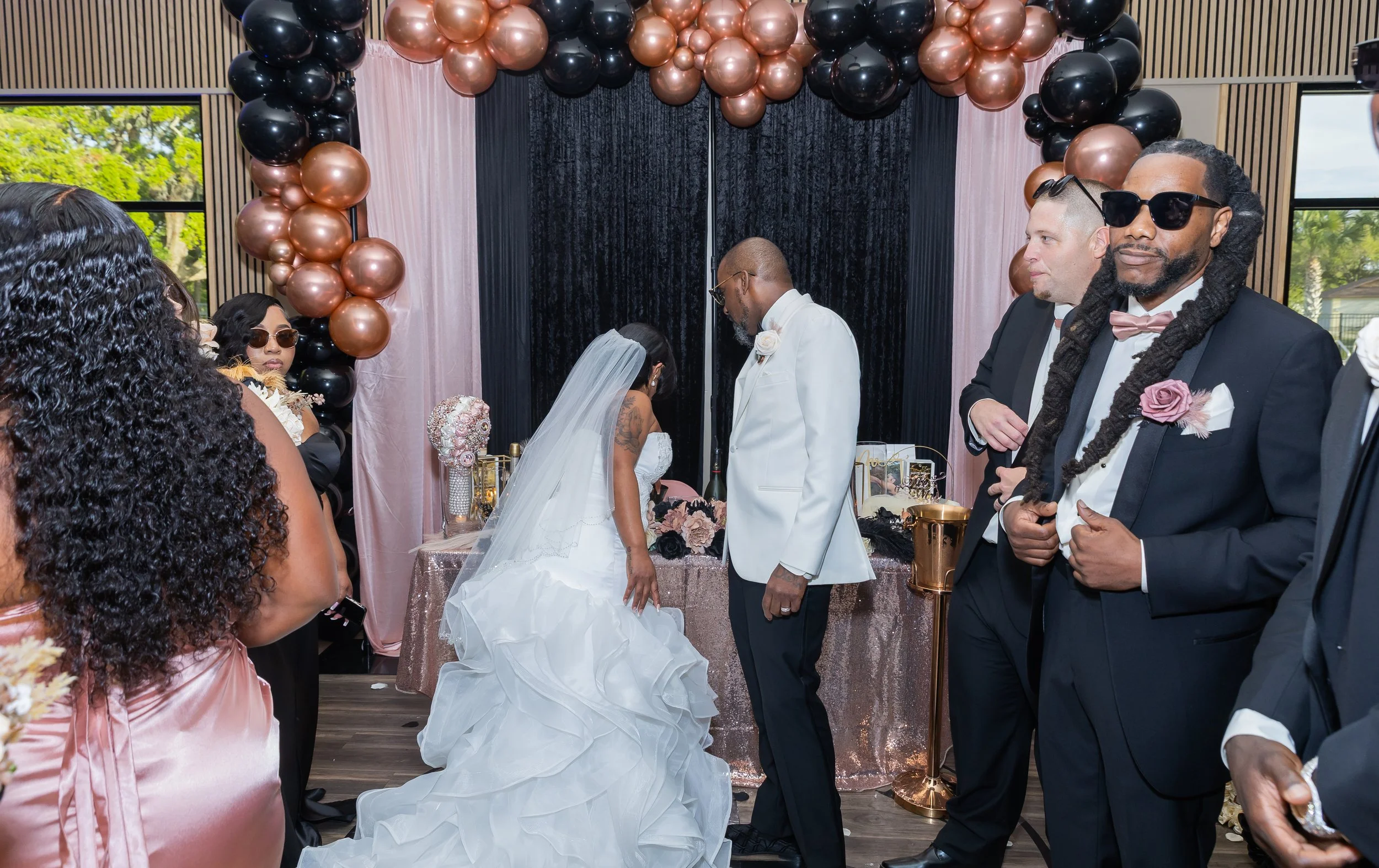 A wedding ceremony with the bride and groom exchanging vows, surrounded by guests, decorated with black and rose gold balloons and elegant drapery.