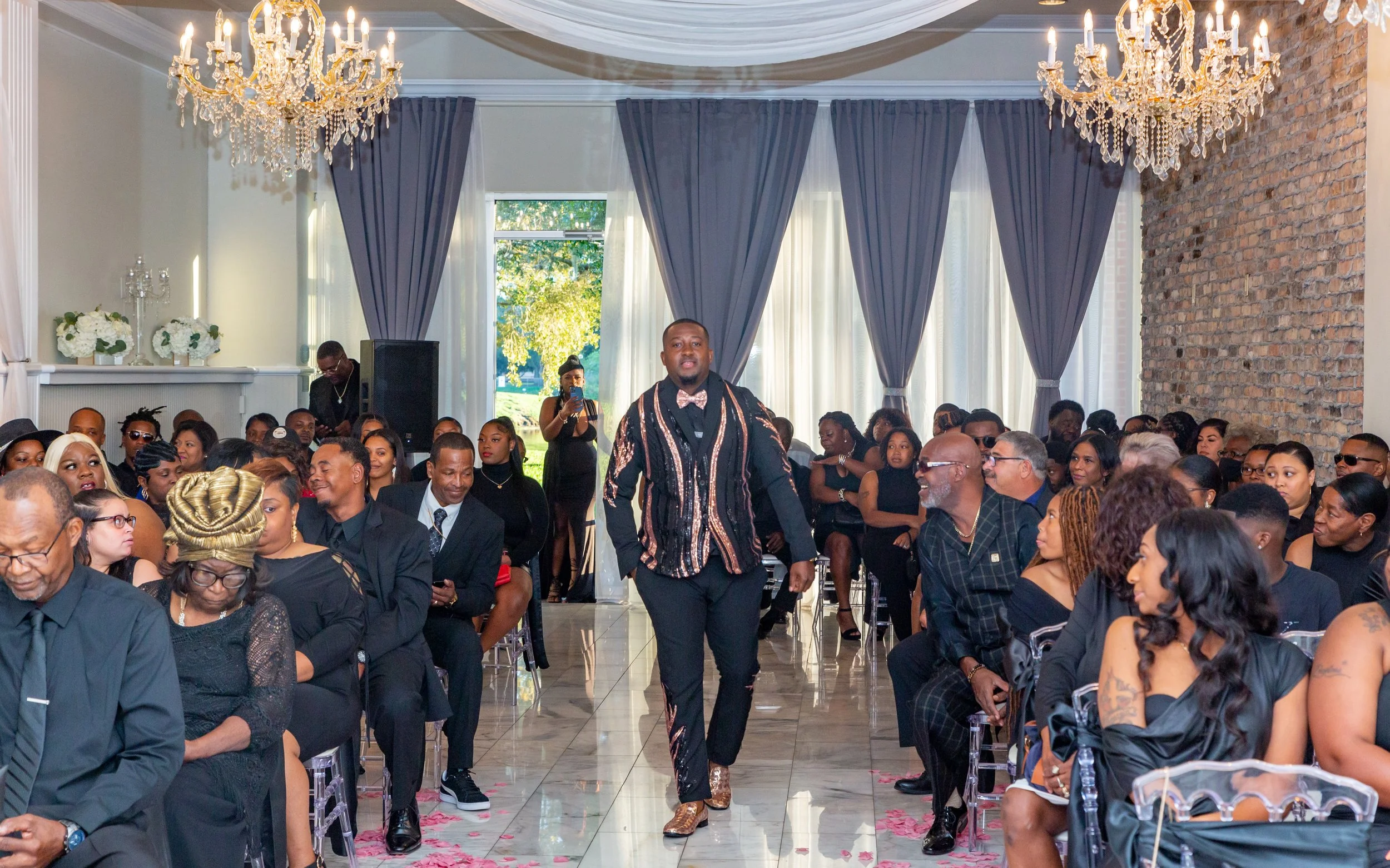 A man walking in front of seated guests at a formal event in a decorated hall with chandeliers, blue curtains, and a brick wall.