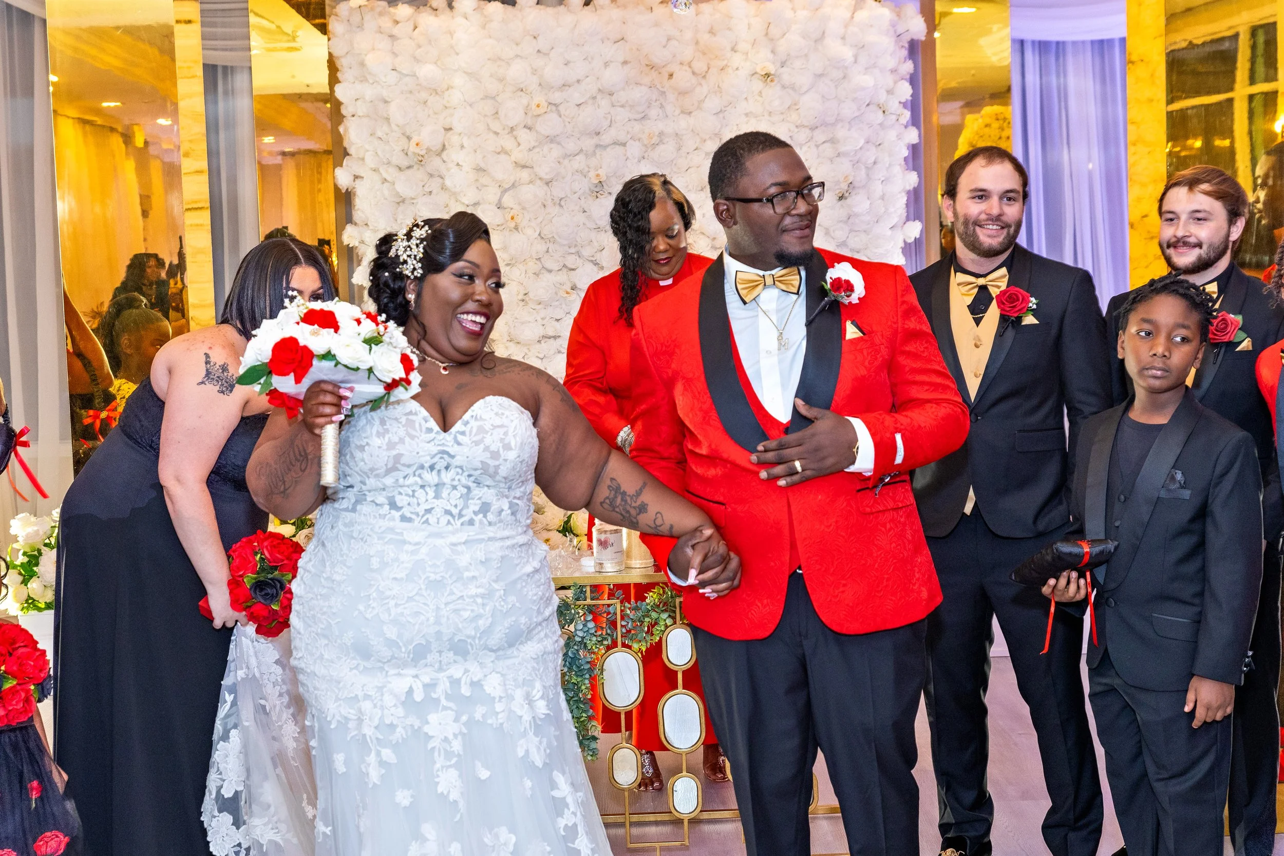 A group of wedding party members, including a bride in a white lace wedding gown holding a bouquet, and the groom wearing a red jacket with gold accents, standing together at a wedding reception with floral and decorative background.