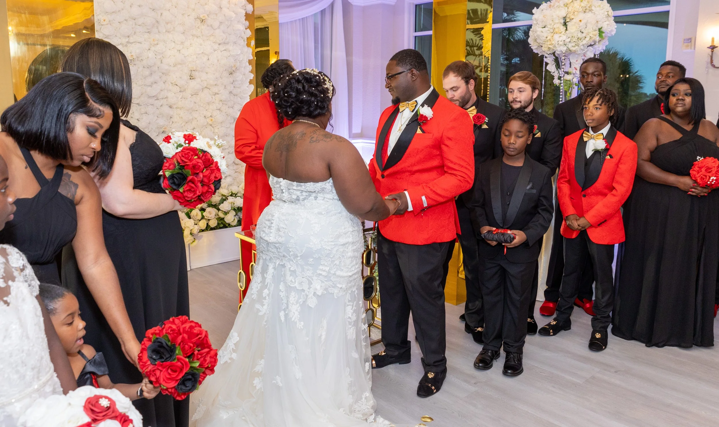 A wedding ceremony with the bride and groom holding hands, surrounded by flower arrangements and family members dressed in formal attire.