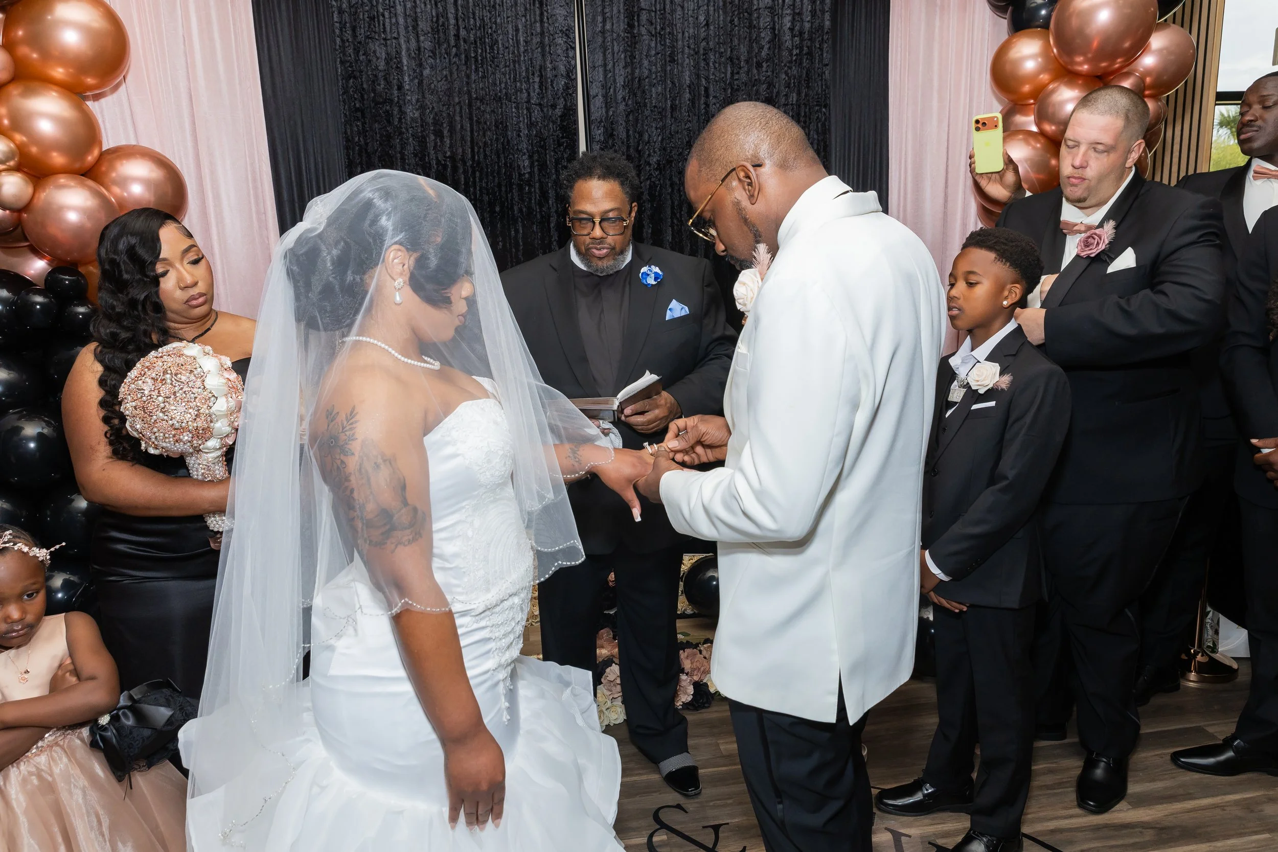 A wedding ceremony with the couple exchanging vows, surrounded by family and friends. The bride is wearing a white wedding dress and veil, and the groom is in a white tuxedo. Bridesmaids and groomsmen are dressed in formal attire, with balloons in bl