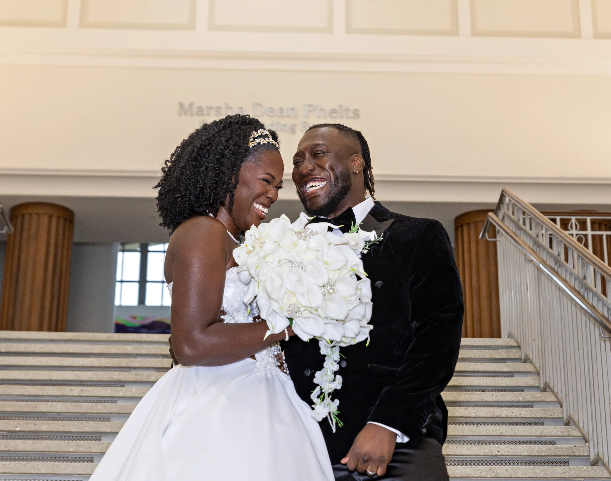 A newlywed couple sharing a joyful moment on a staircase inside a building, with the bride in a white wedding dress and the groom in a black tuxedo, holding a bouquet of white orchids.